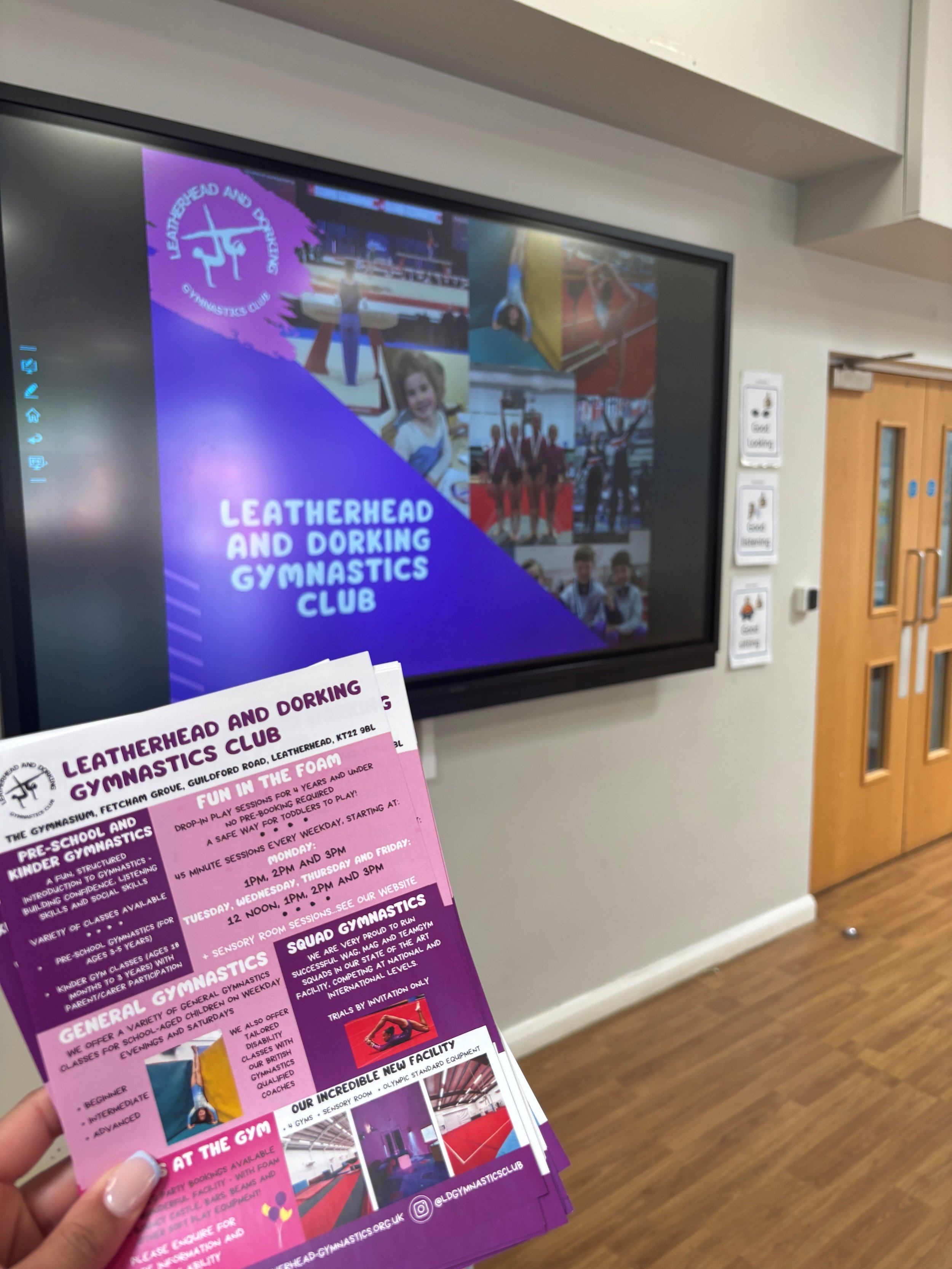 A person holding a flyer or brochure for the Leatherhead and Dorking Gymnastics Club. The flyer contains information about various gymnastics classes, including pre-school, squad gymnastics, and general gymnastics, with images of gymnasts and the club's contact details. The background shows a digital display screen mounted on a wall inside a building with wooden doors nearby.