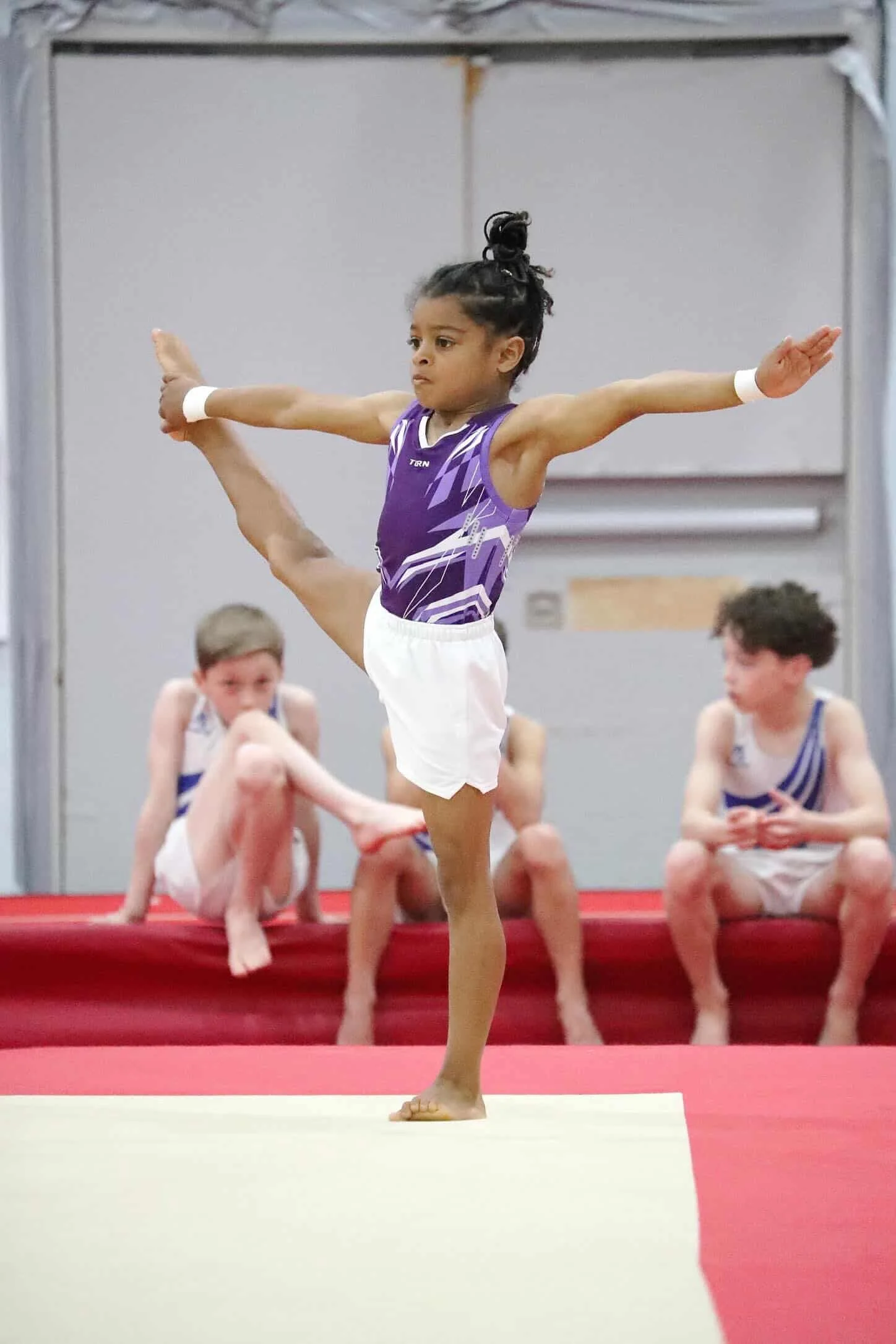 Young girl in a purple gymnastics uniform balancing on her right leg with her left leg extended behind her, holding her foot with her left hand, surrounded by other children in similar uniforms on a gymnastics mat.