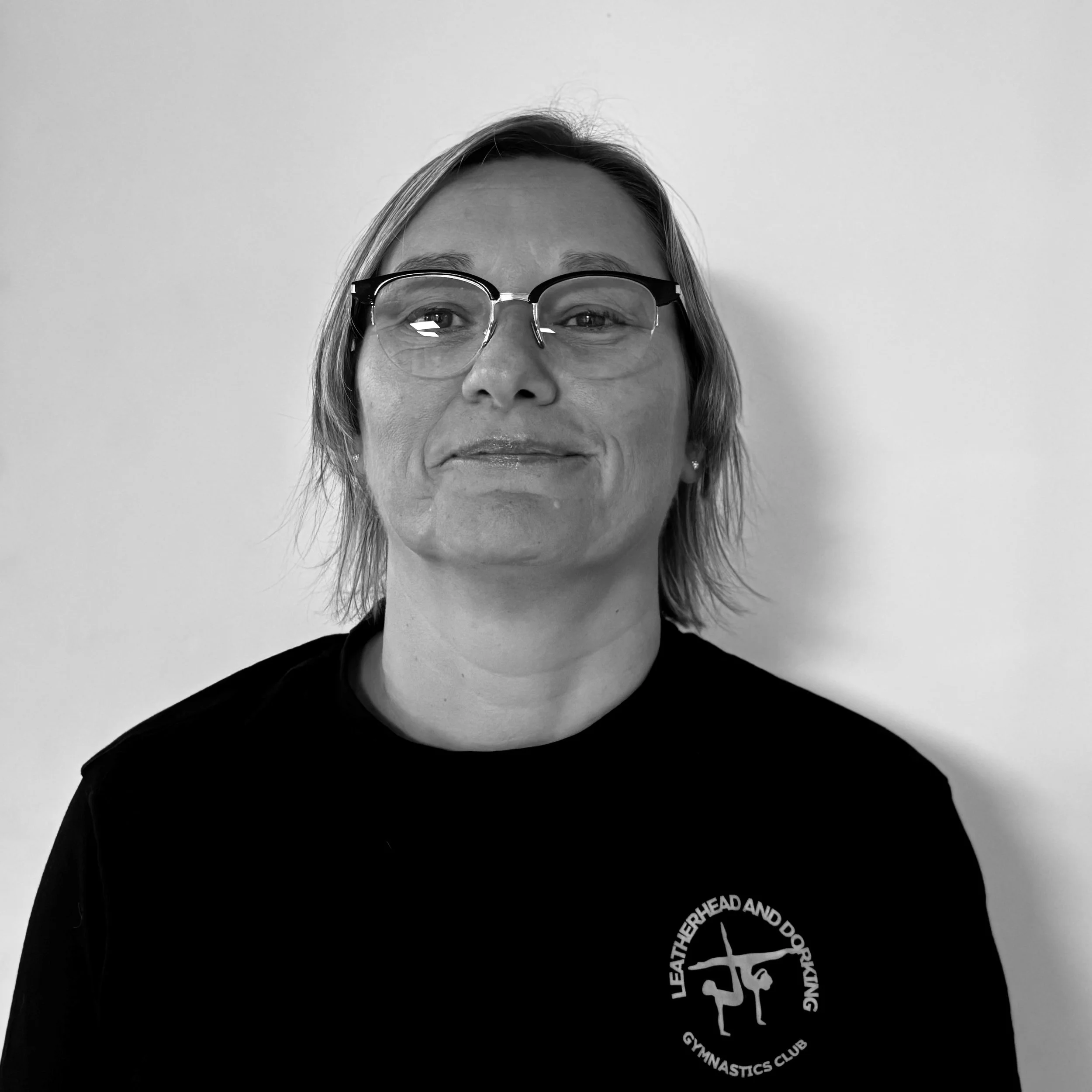 Close-up of a woman with glasses and short hair, smiling slightly, against a plain light background. She wears a black shirt with a logo on the front.