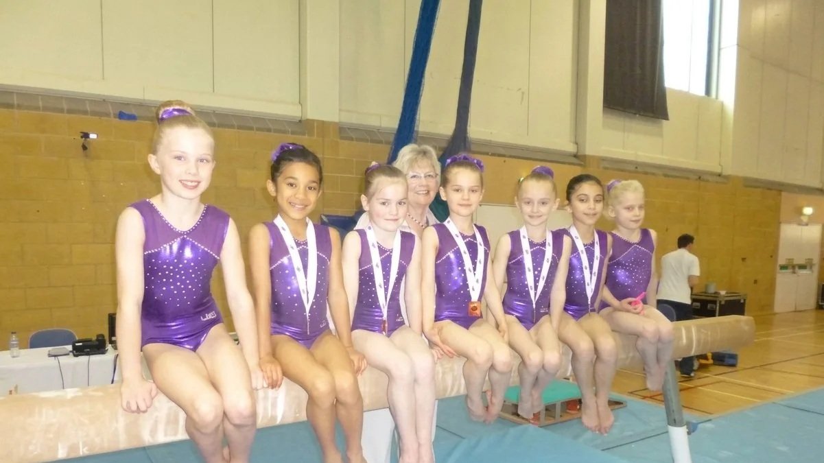 Seven young girls in purple gymnastics leotards sitting with a coach behind a gymnastics beam in a gymnasium, all wearing medals.