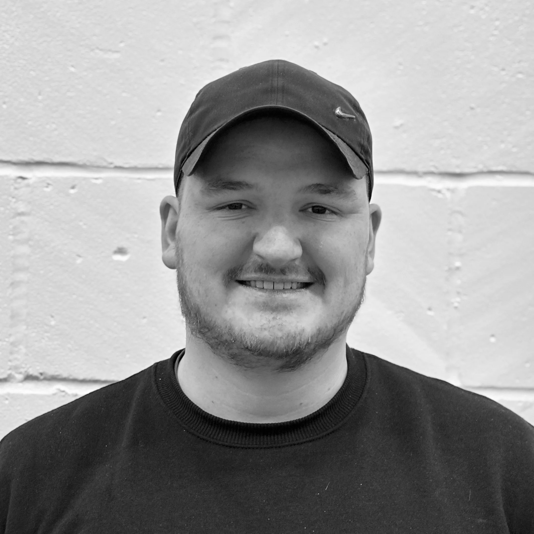 Black and white photo of a smiling young man wearing a dark baseball cap and a black crewneck shirt, with a textured wall in the background.