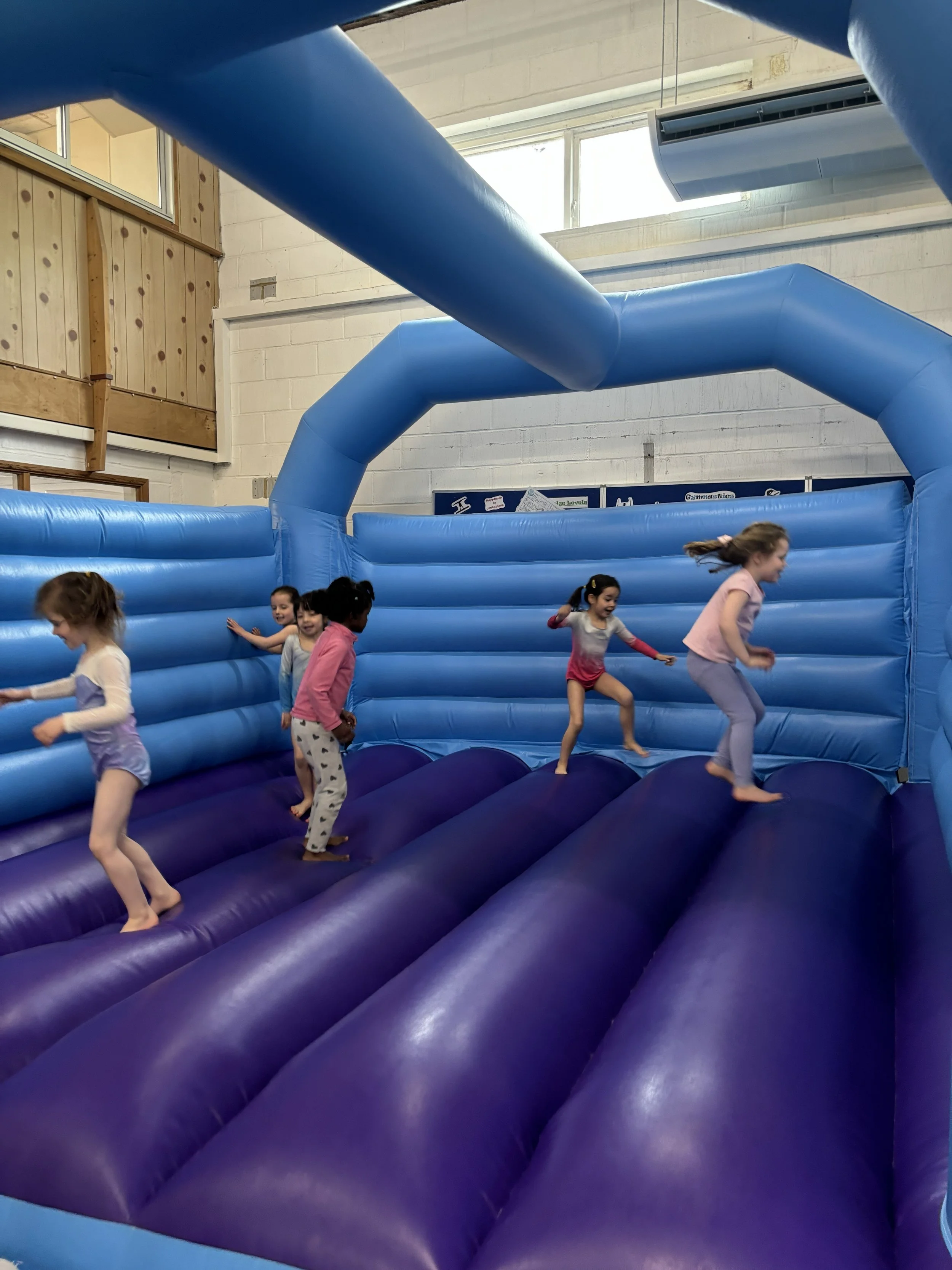 Children playing on an inflatable bounce house with blue walls, located indoors with white brick walls and windows.
