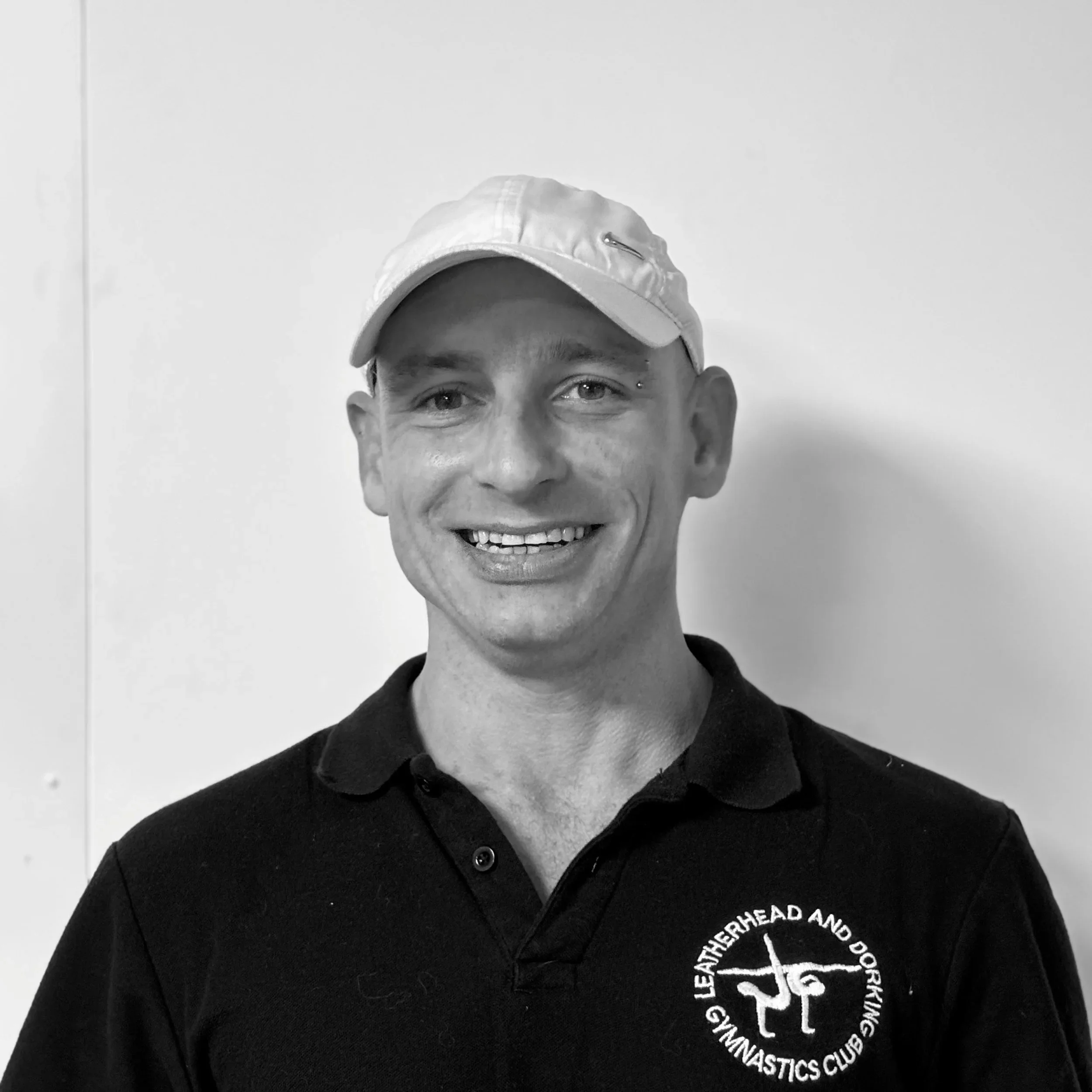 Close-up of a smiling person's face wearing a baseball cap and a black shirt with a logo of a gymnastics club. The person's eyes are visible, and they have a dermal piercing above their right eyebrow.
