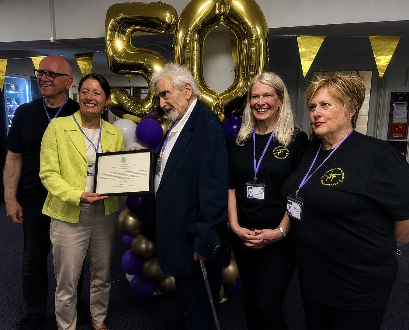 Group of five people celebrating, with large gold number balloons reading '50' and purple and white balloons behind them. The woman second from the left is holding a framed certificate. They are smiling and wearing casual to semi-formal attire, with some having lanyards and name tags.
