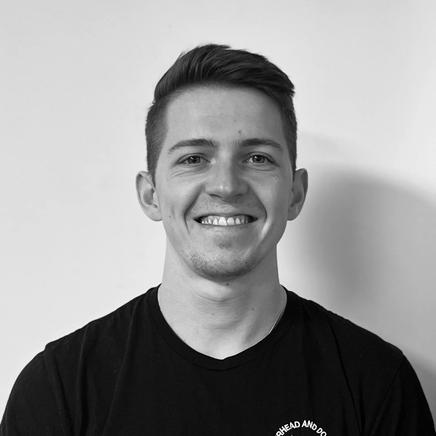 A young man with short dark hair, smiling, wearing a black shirt, standing against a plain white wall.