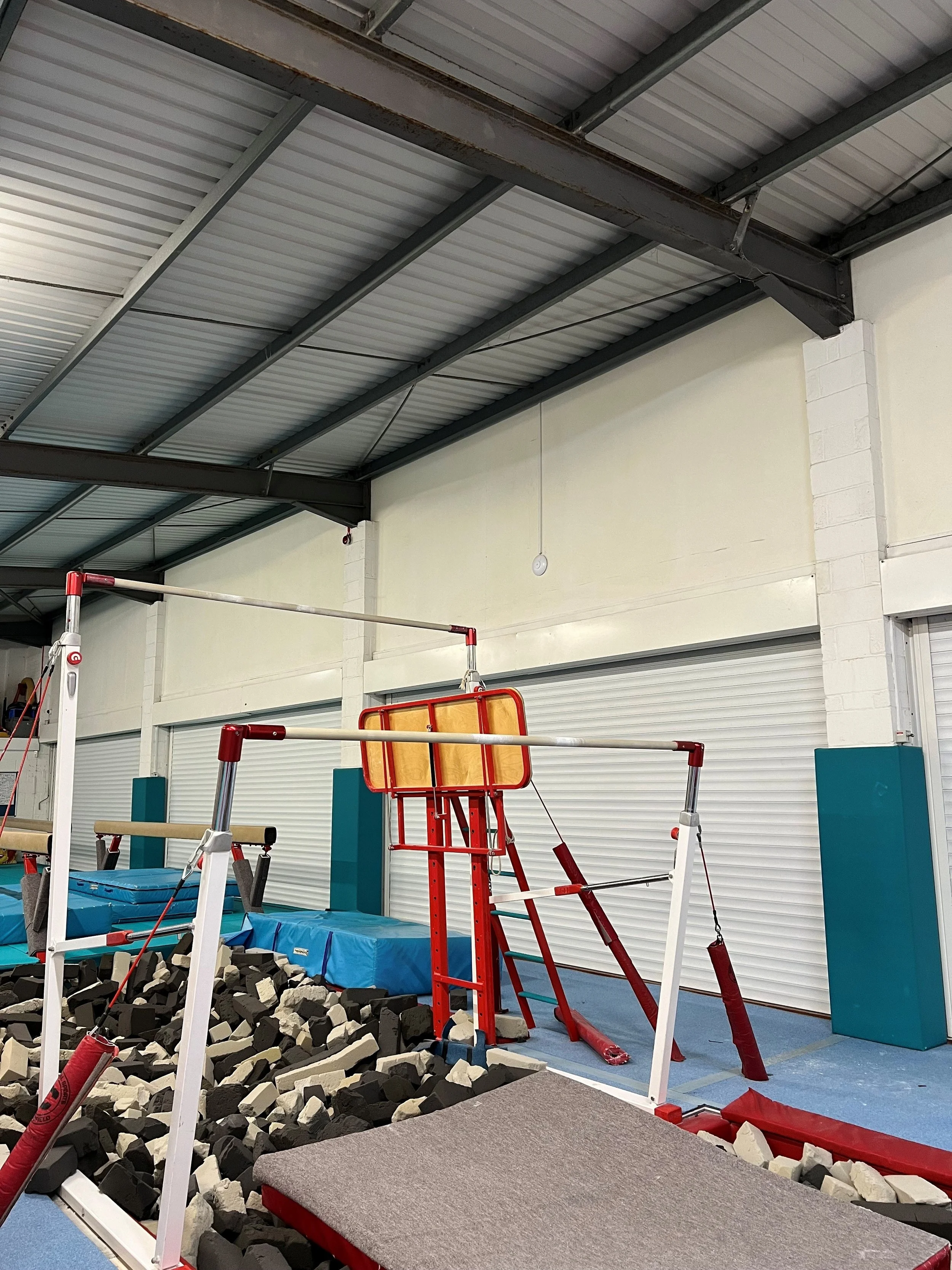 An indoor gym with gymnastics equipment, including a foam pit filled with black and white foam blocks, balance beams, and a trampoline, under a metal roof ceiling.
