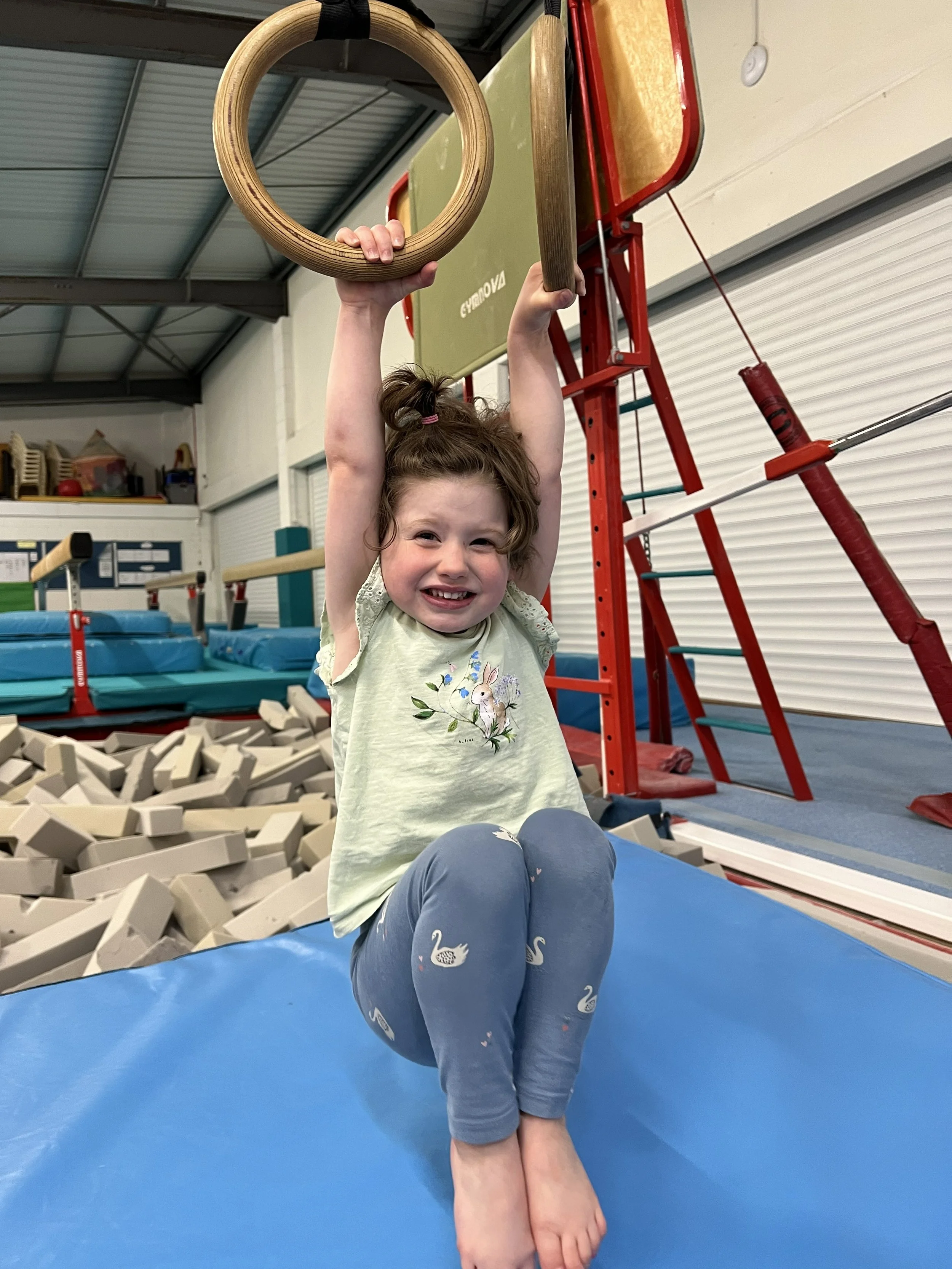 A young girl sitting on a blue gym mat, smiling, with her legs folded beneath her. She is holding onto gymnastic rings hanging from the ceiling in a gym or indoor athletic facility, with foam blocks on the floor and gym equipment visible in the backg