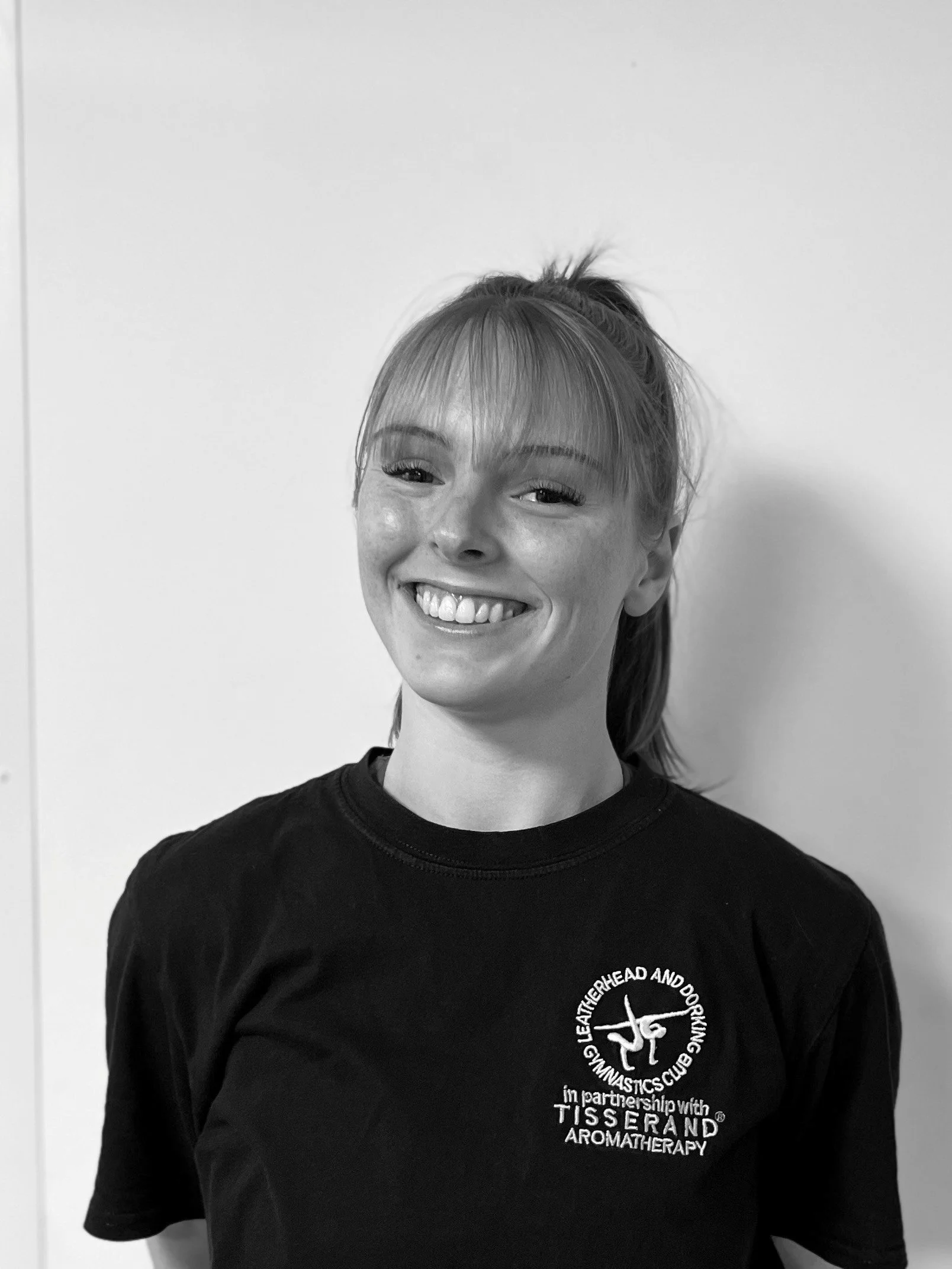 A woman with light skin and straight hair tied back, smiling, wearing a black T-shirt with a logo and text related to gymnastics and aromatherapy, standing in front of a plain white background.