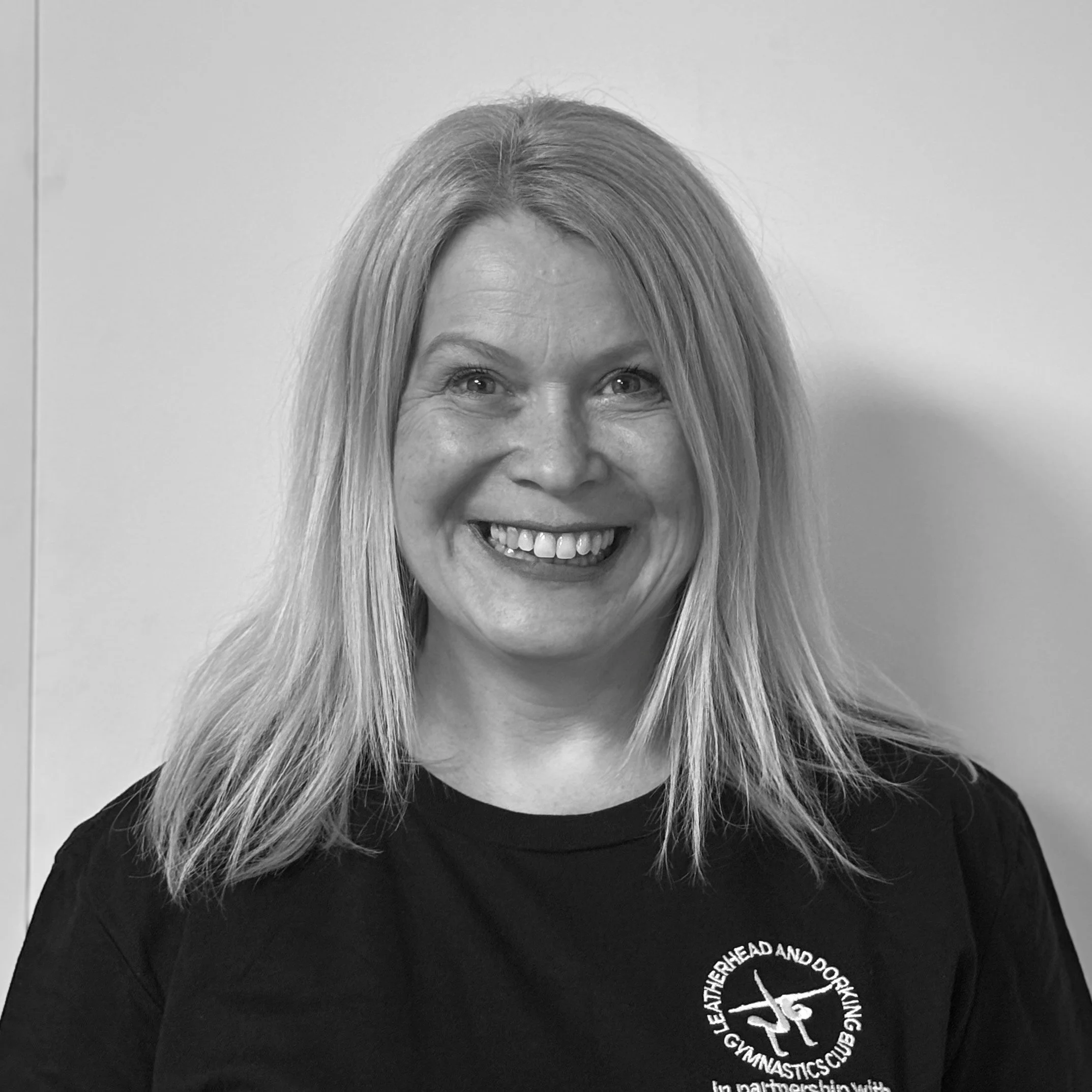 Close-up of a woman's face with blonde hair, smiling, wearing a black t-shirt with white text and a logo.