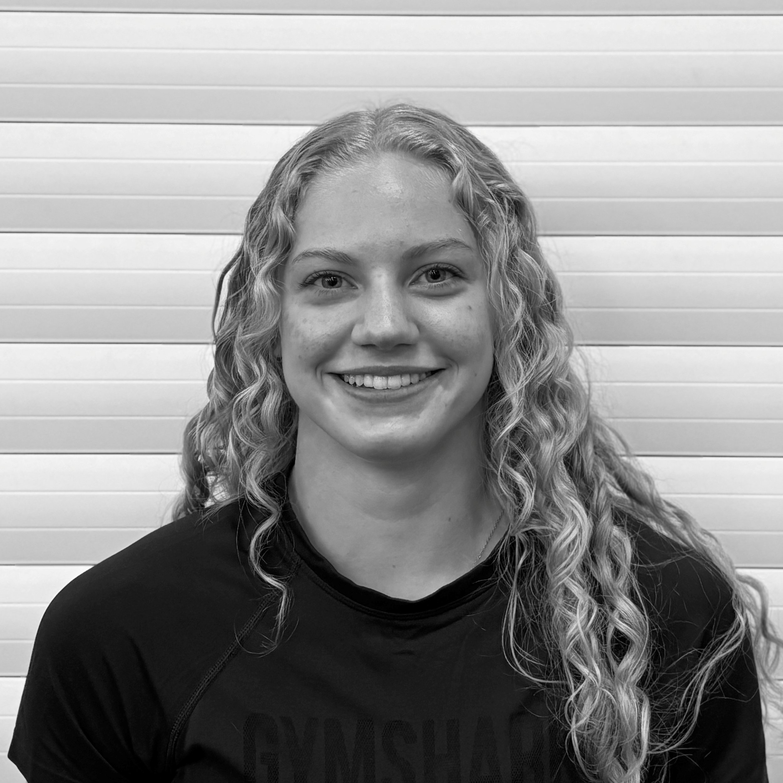 Close-up of a woman with curly hair smiling, wearing a black top and standing in front of a horizontal-striped wall.