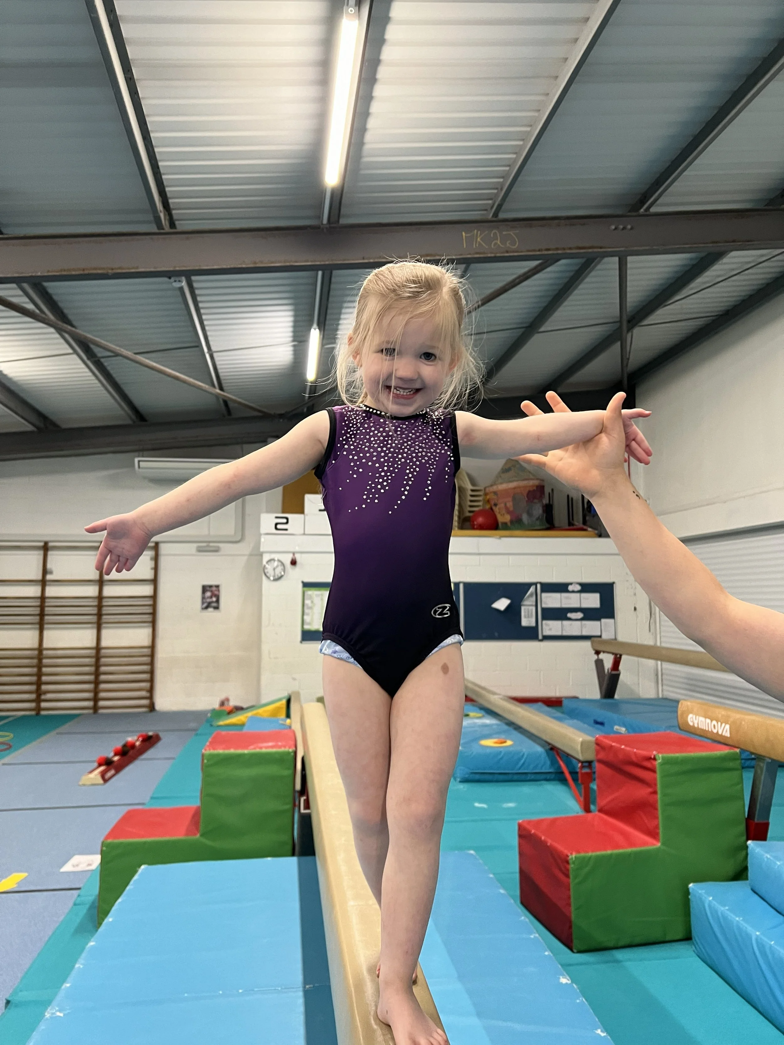 A young girl in a purple gymnastics leotard is balancing on a thin wooden balance beam in an indoor gym. She is smiling, and an adult's hand is supporting her from the side. The background shows gymnastic equipment, pads, and a blue floor mat.