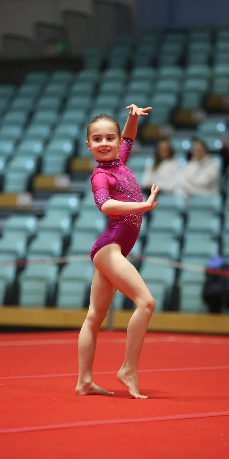 A young girl in a pink gymnastics leotard performing a routine on a red mat in an indoor arena, with empty blue spectator seats in the background.