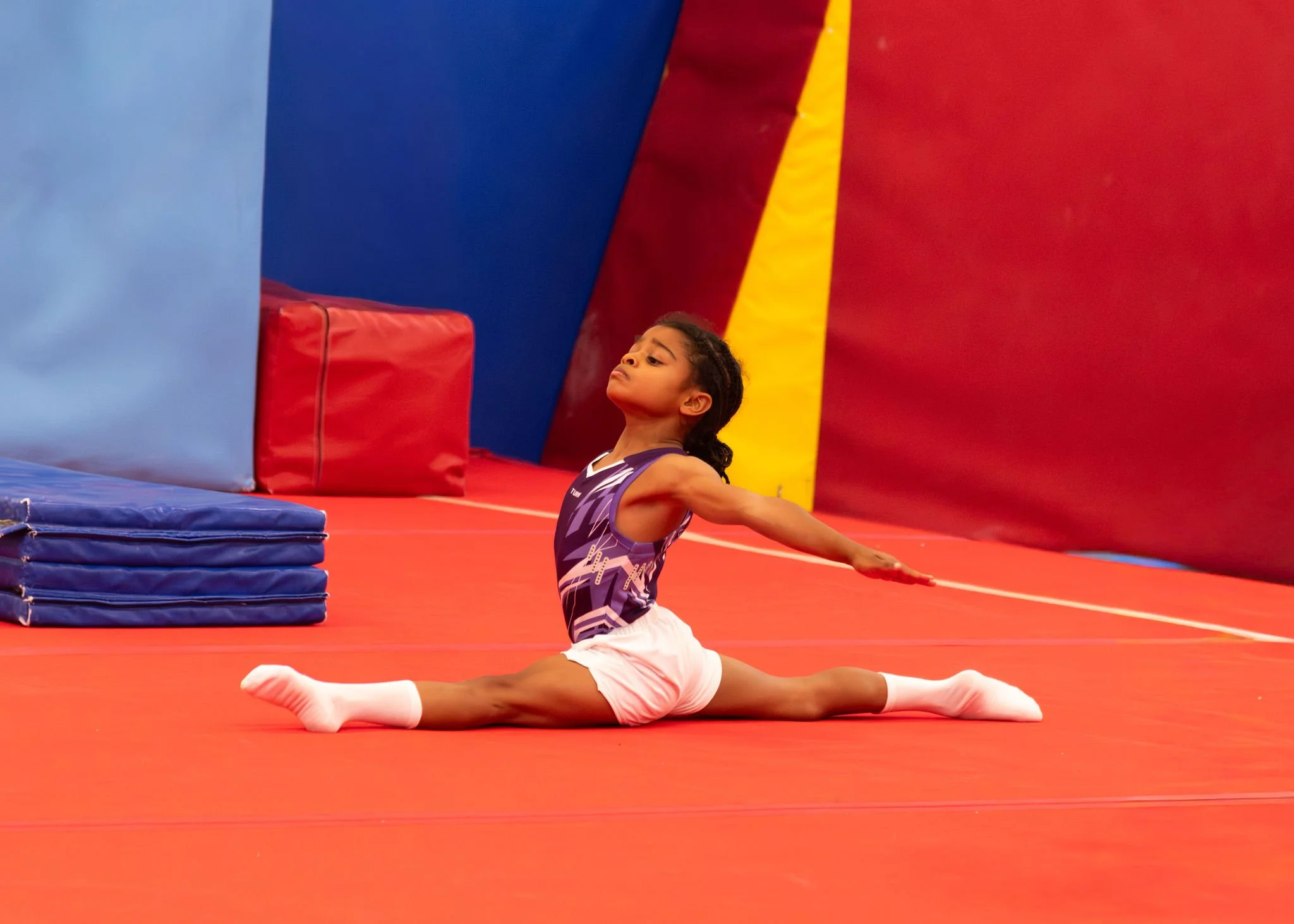 A young boy performing a split stretch on a red gymnastics mat inside a colorful gymnastics training facility with padded walls and stacked mats in the background.