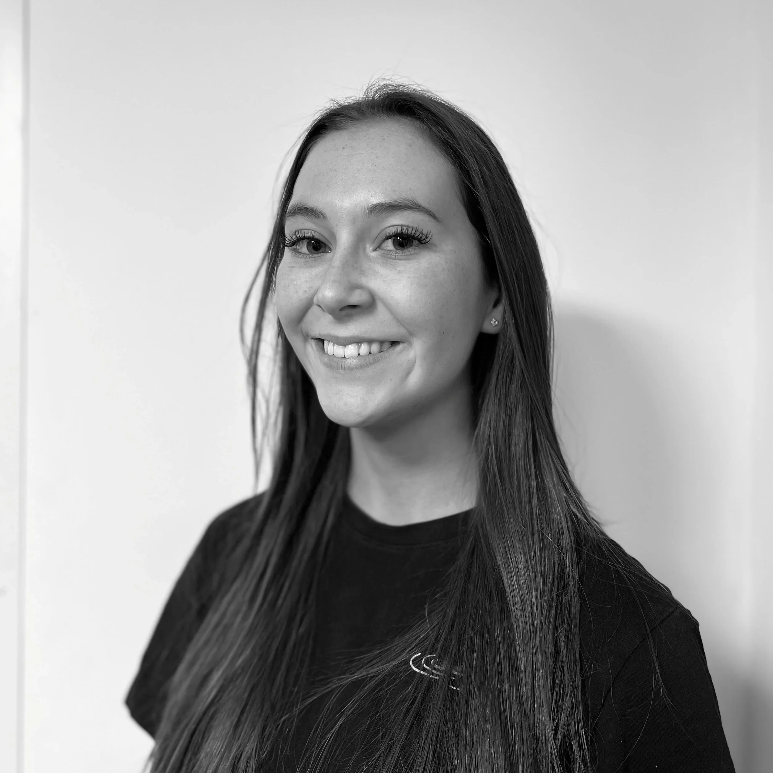 A young woman with long dark hair, smiling, showing braces, standing against a plain white background.