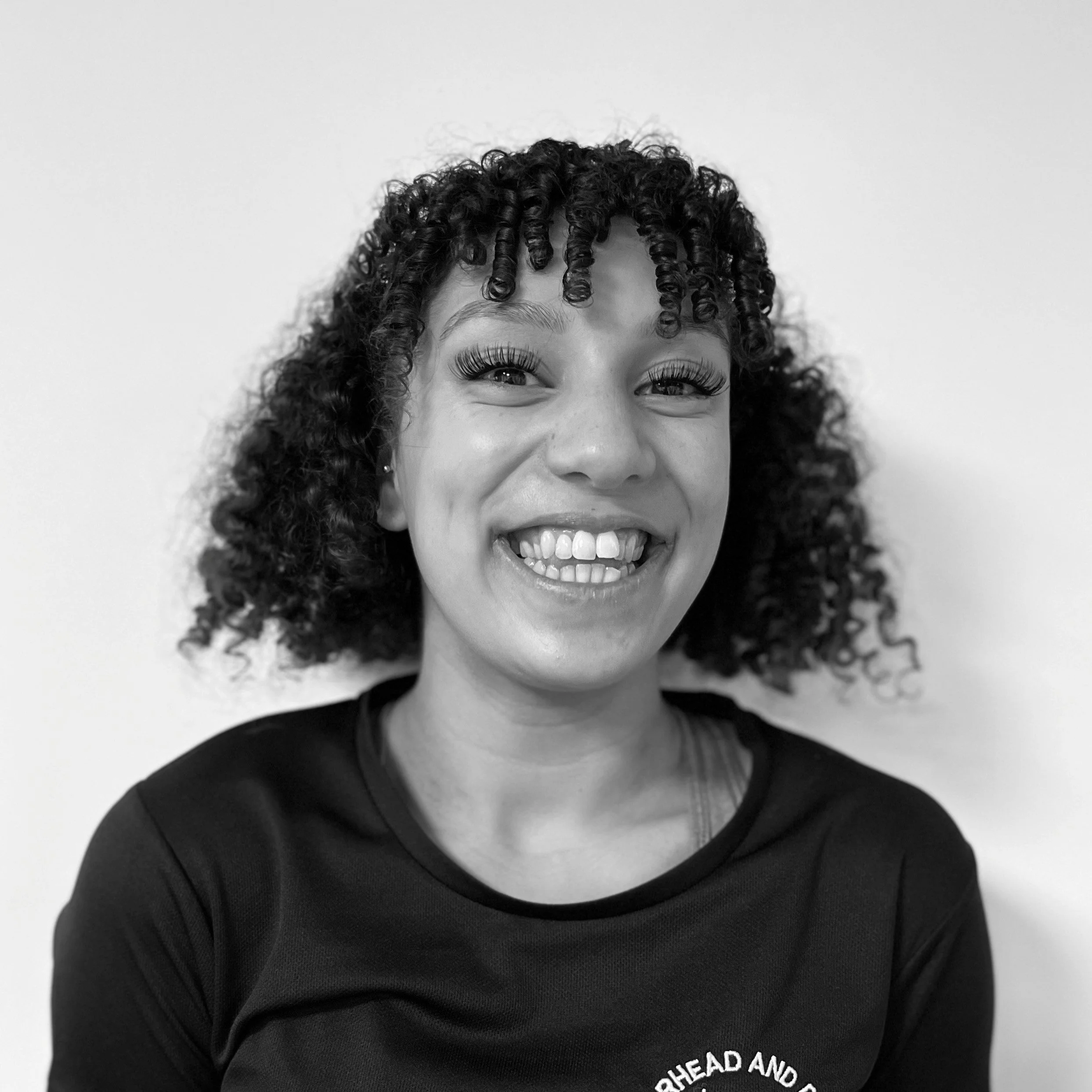 Close-up of a smiling woman with curly hair, wearing a black shirt with partial text visible, against a plain background.