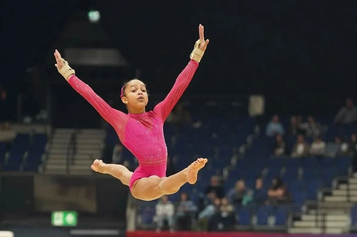 Young girl performing a gymnastics leap in a pink outfit with a high leap in an indoor arena with spectators.