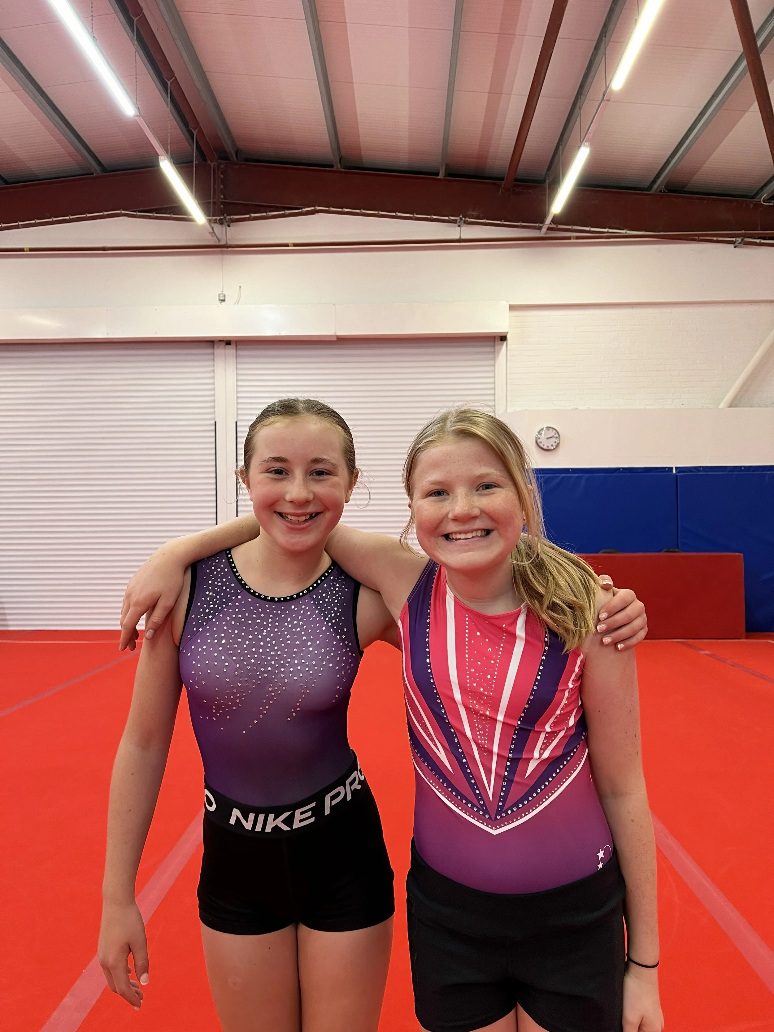 Two young female gymnasts standing side by side, smiling, with their arms around each other's shoulders, in a gymnasium. Both are wearing athletic leotards, one in purple with white dots and the other in pink with white and purple design, with a foam