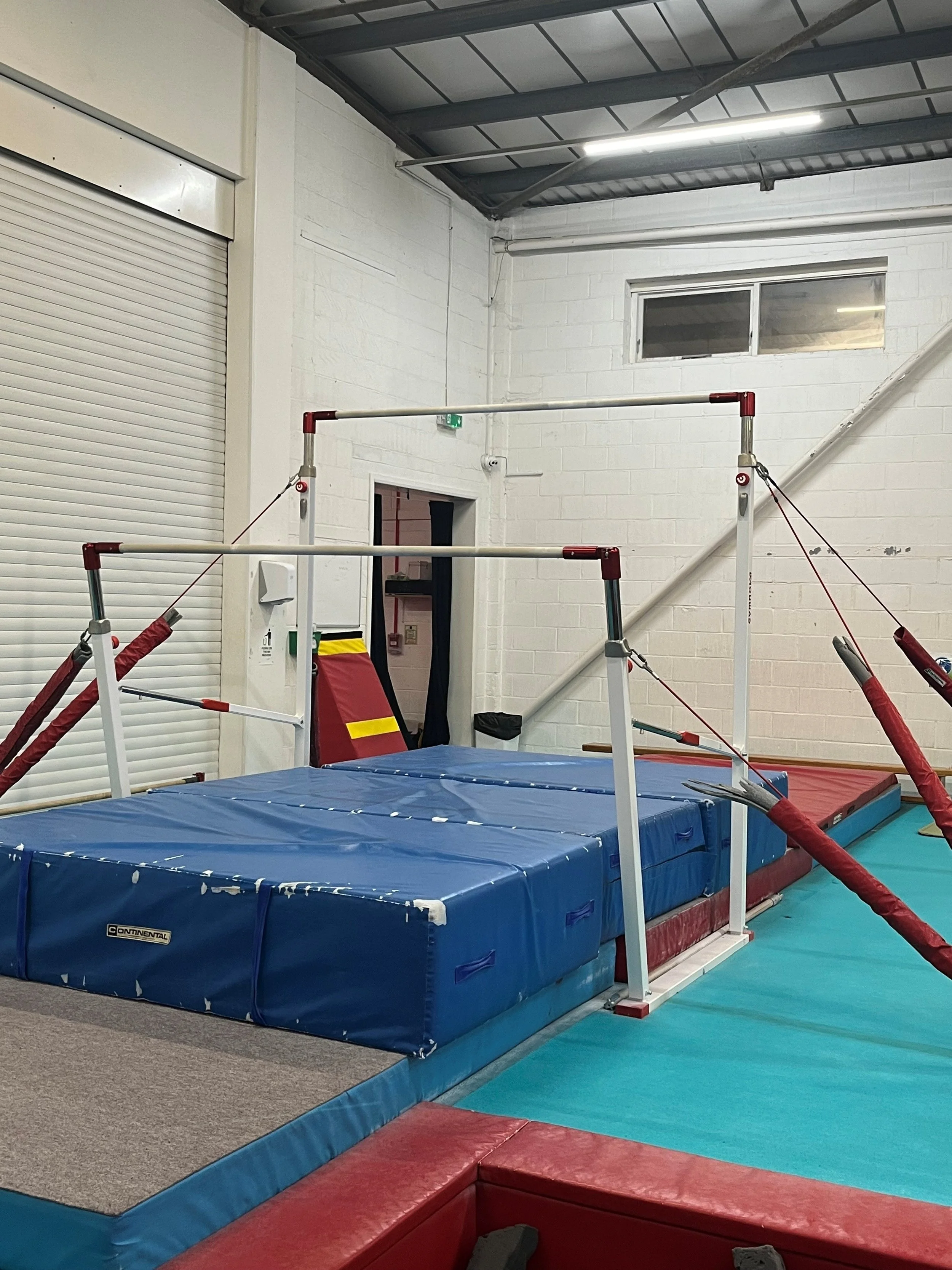 Indoor gymnasium with gymnastics equipment including a padded vaulting box, a springboard, and a high bar. The walls are white with a small window near the ceiling, and there is a closed garage door on the left. The flooring is a mix of mats and foam padding.