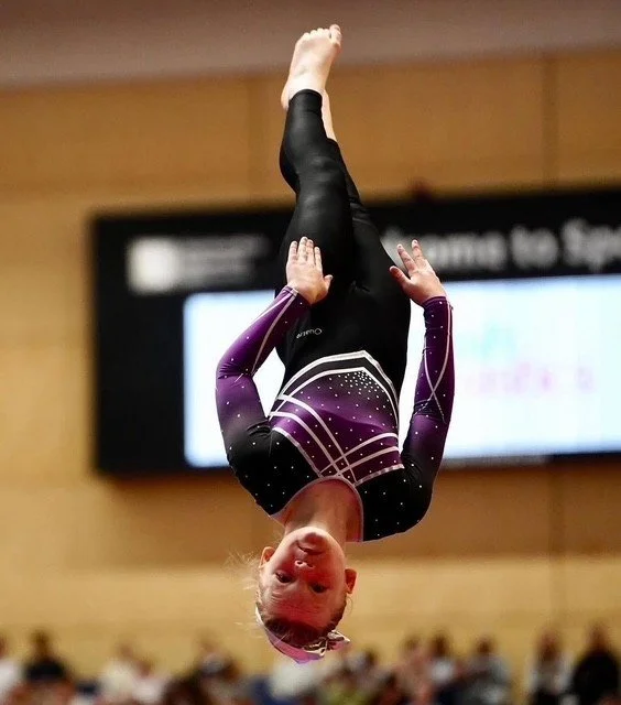 A young gymnast performing an upside-down pose in a gymnasium, wearing a purple and black leotard with white accents.