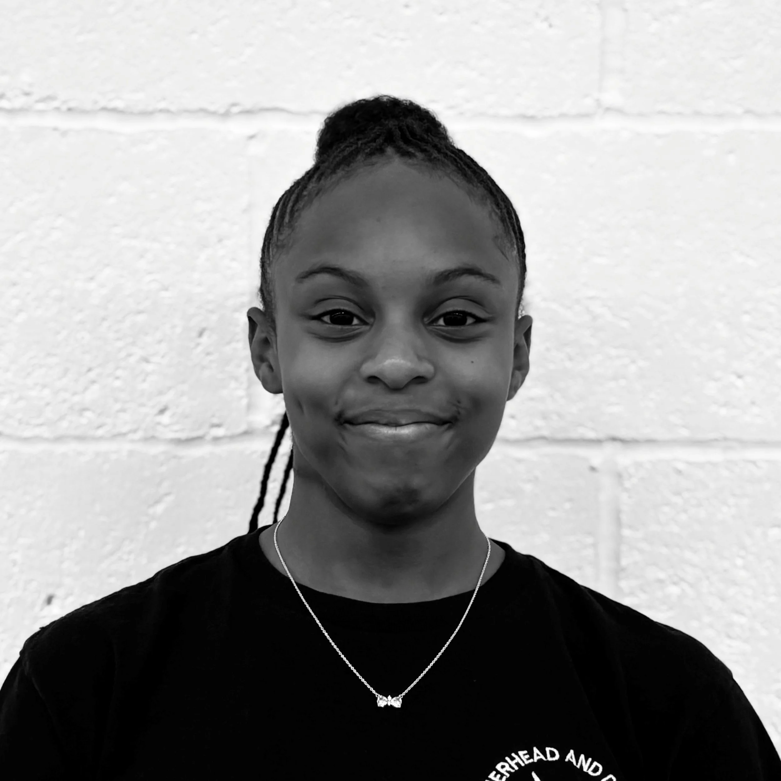 Close-up of a young girl smiling, wearing a black sweatshirt with a circular logo and a necklace with a butterfly pendant, standing in front of a white brick wall.