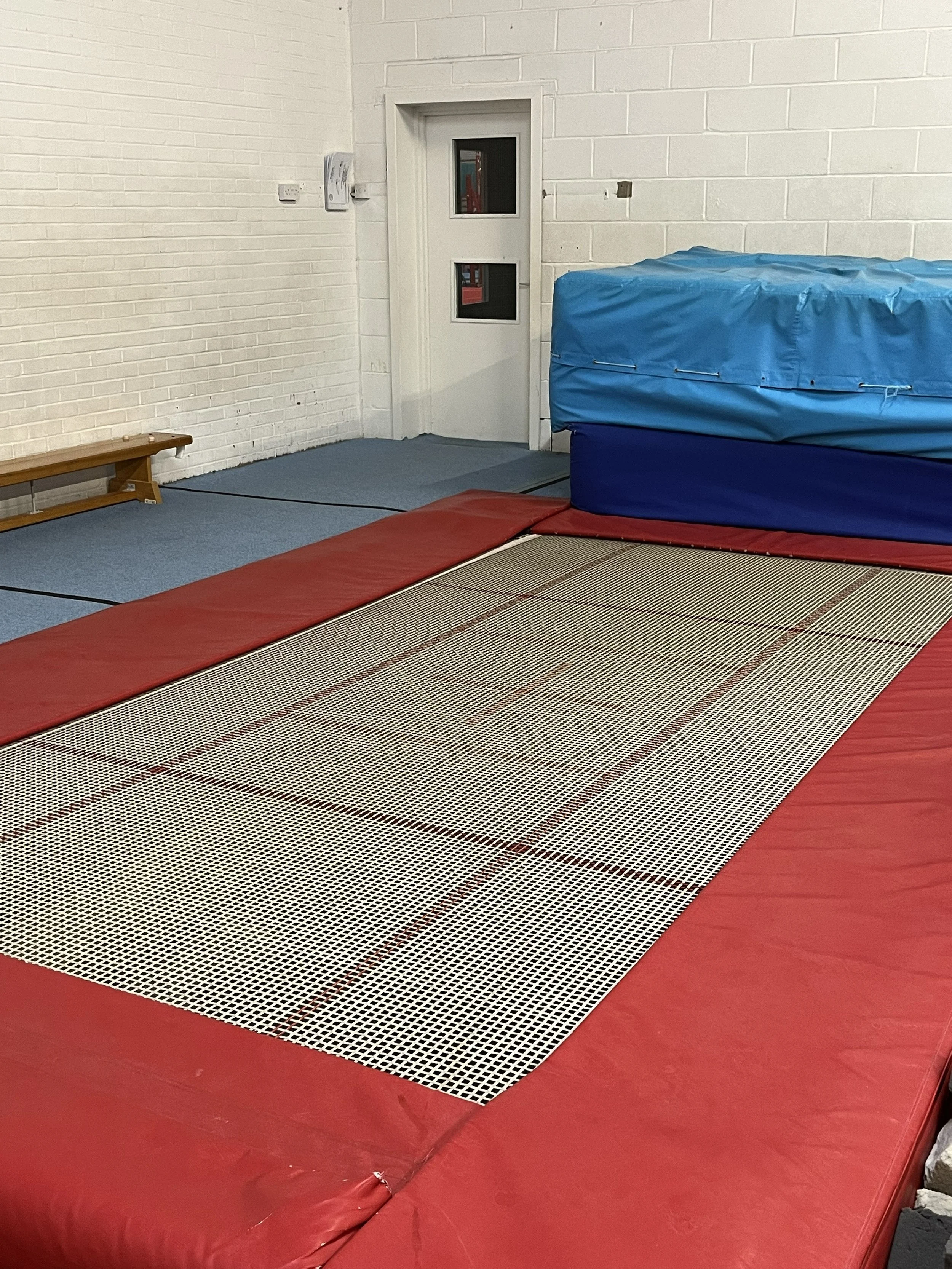 Gymnasium with red mats, a springboard, a blue padded vault, and a white brick wall.