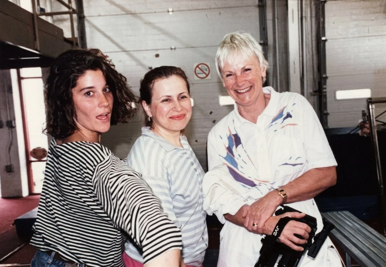 Three women standing together in a garage or warehouse-like space, smiling and posing for the photo. The woman on the left has curly dark hair, the woman in the middle has straight brown hair, and the woman on the right has short white hair and is holding a camera.