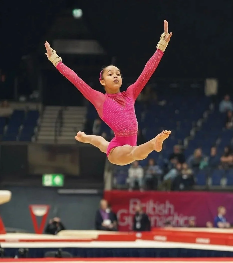 A young gymnast in a pink leotard mid-air performing on the balance beam at a gymnastics competition.
