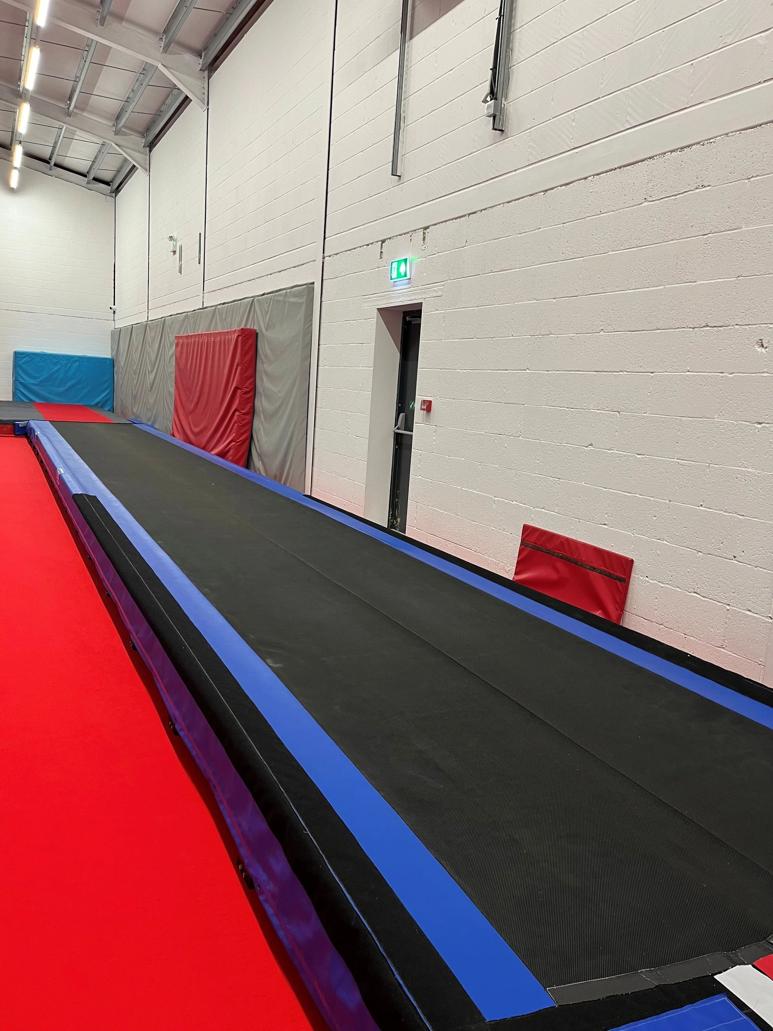 Indoor trampoline with padded surrounding mats and a bright red floor in a gym or recreation center.