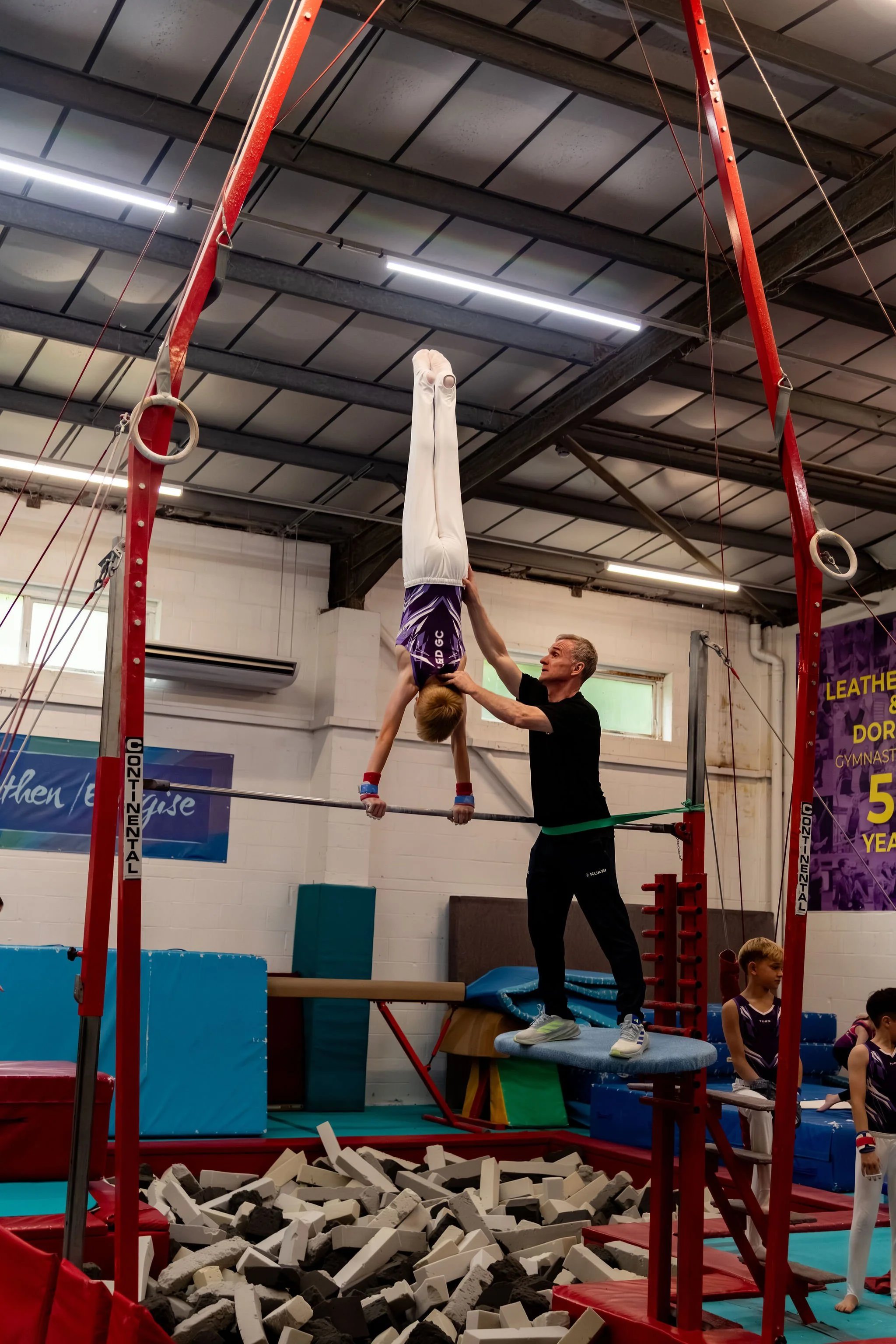 A gymnast practicing on the high bar assisted by a coach in a gymnasium. The gymnast is upside down with hands gripping the bar, while the coach supports her from below. Foam blocks are on the ground beneath the equipment.