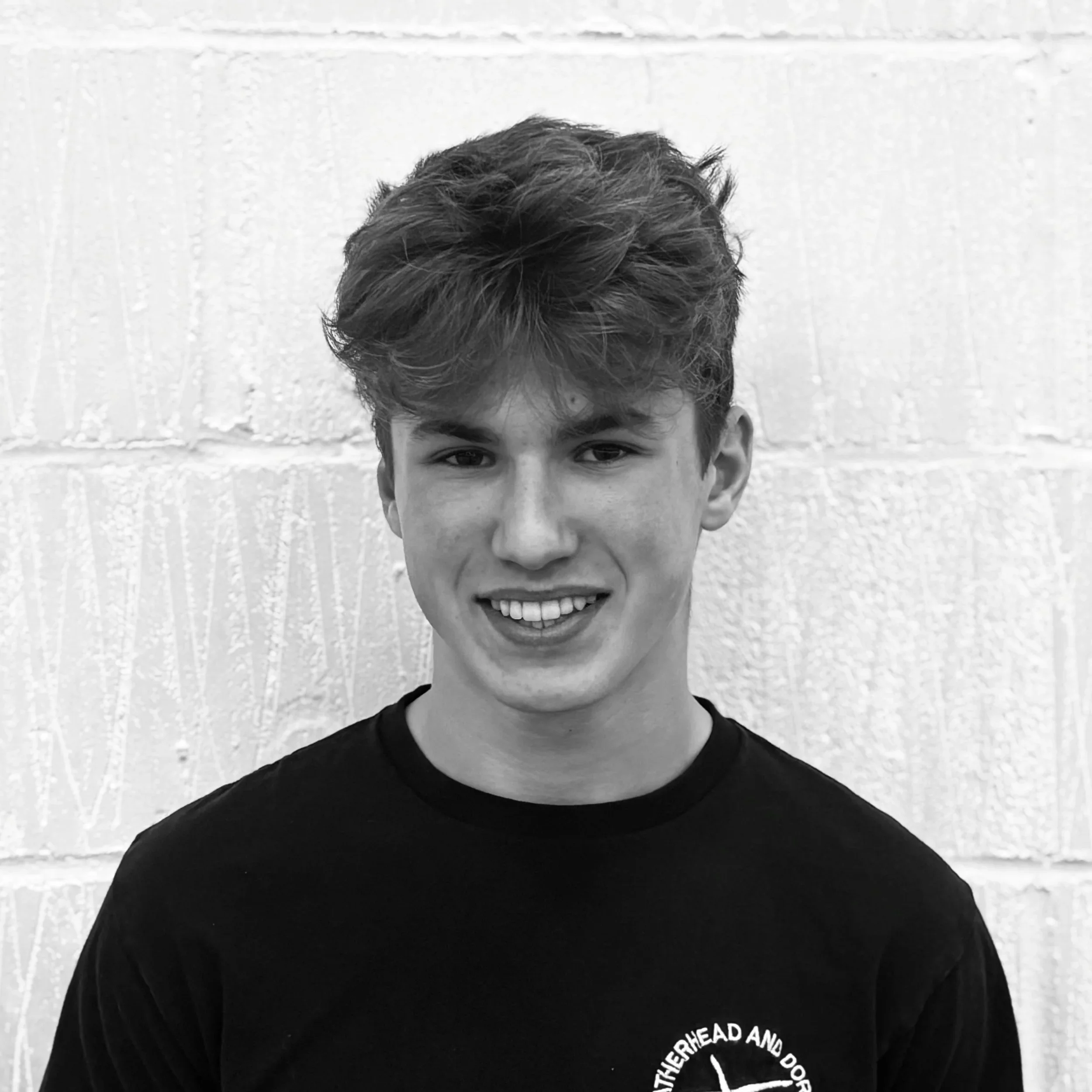 Close-up of a young man's face with curly hair, smiling, against a light-colored textured wall background.