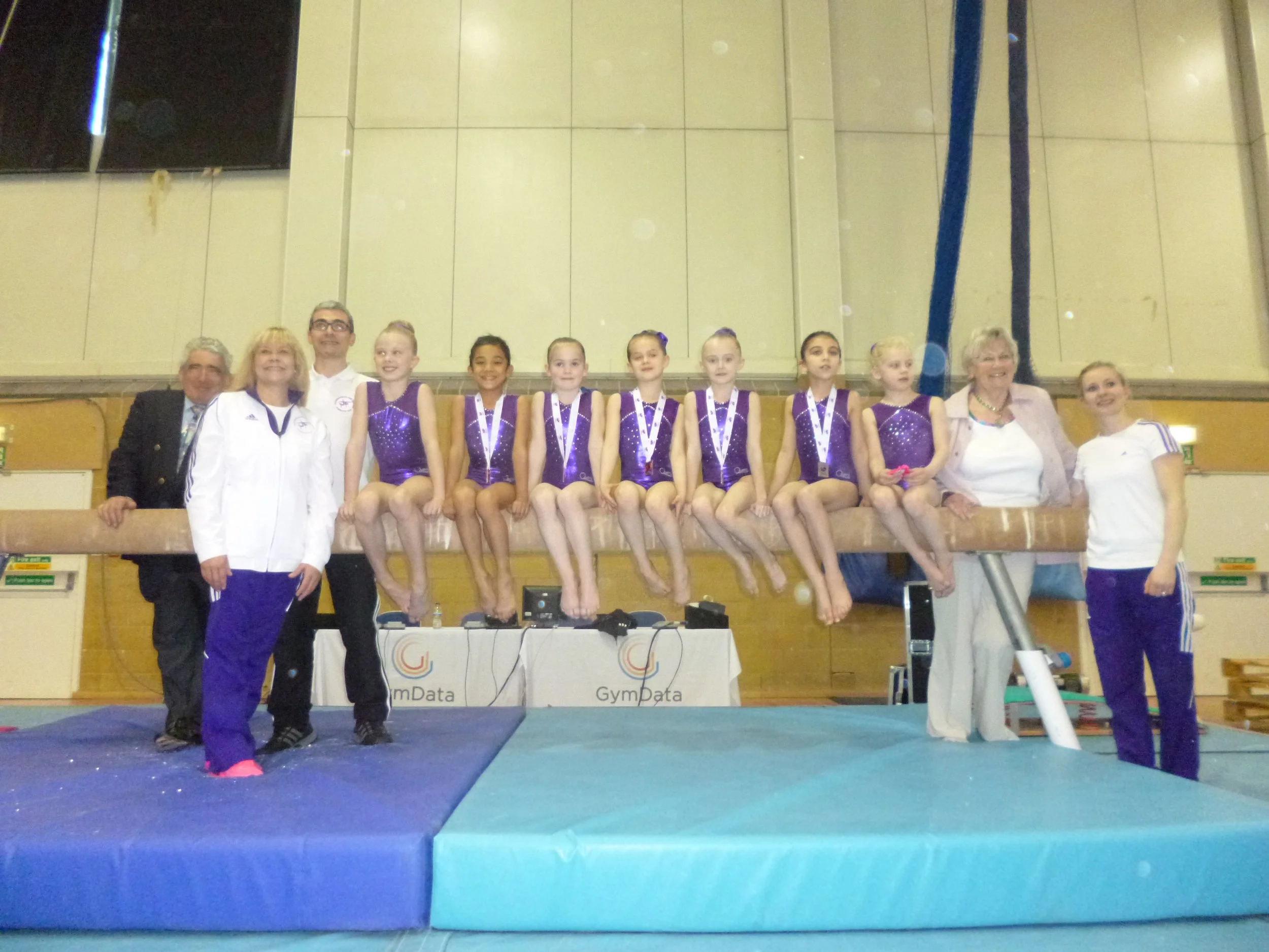 Group of young gymnasts wearing purple leotards and medals, sitting on a balance beam with officials and coaches standing behind them, in a gymnasium.