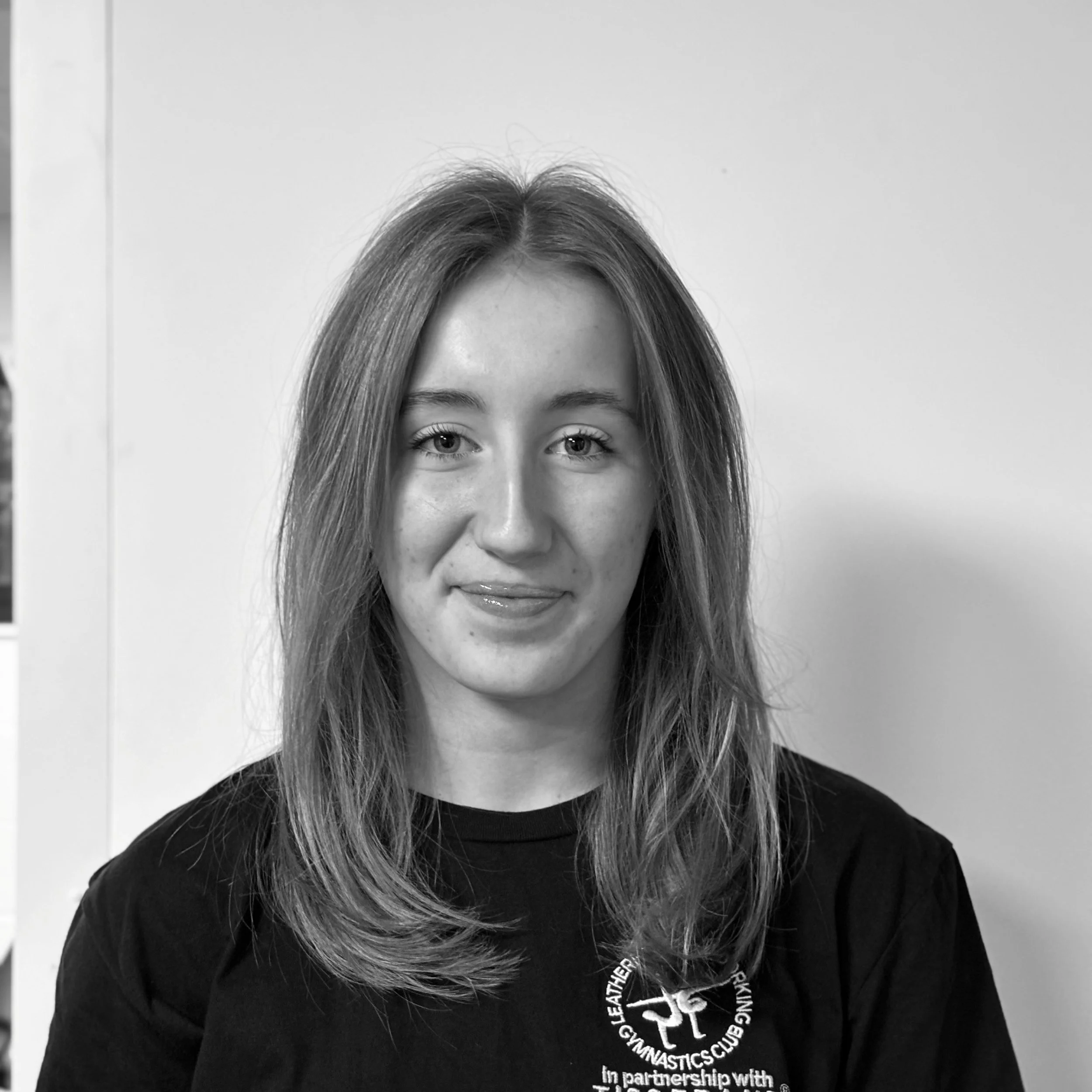 A young woman with long hair and a slight smile, wearing a black shirt with white text and a logo, standing against a plain light-colored wall.