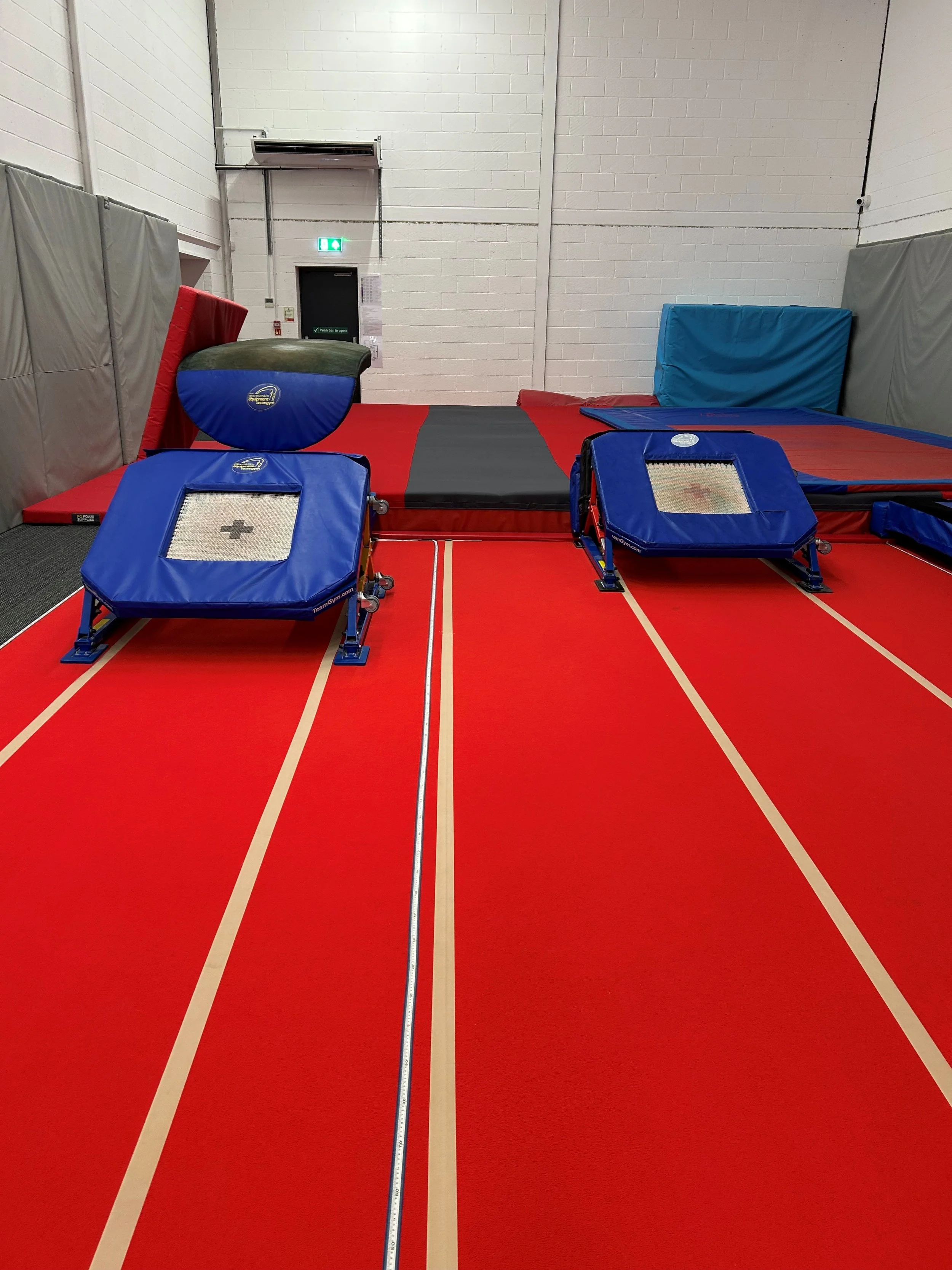 Indoor gym with red and black mats, blue padded obstacles, and foam cushions for gymnastics training.
