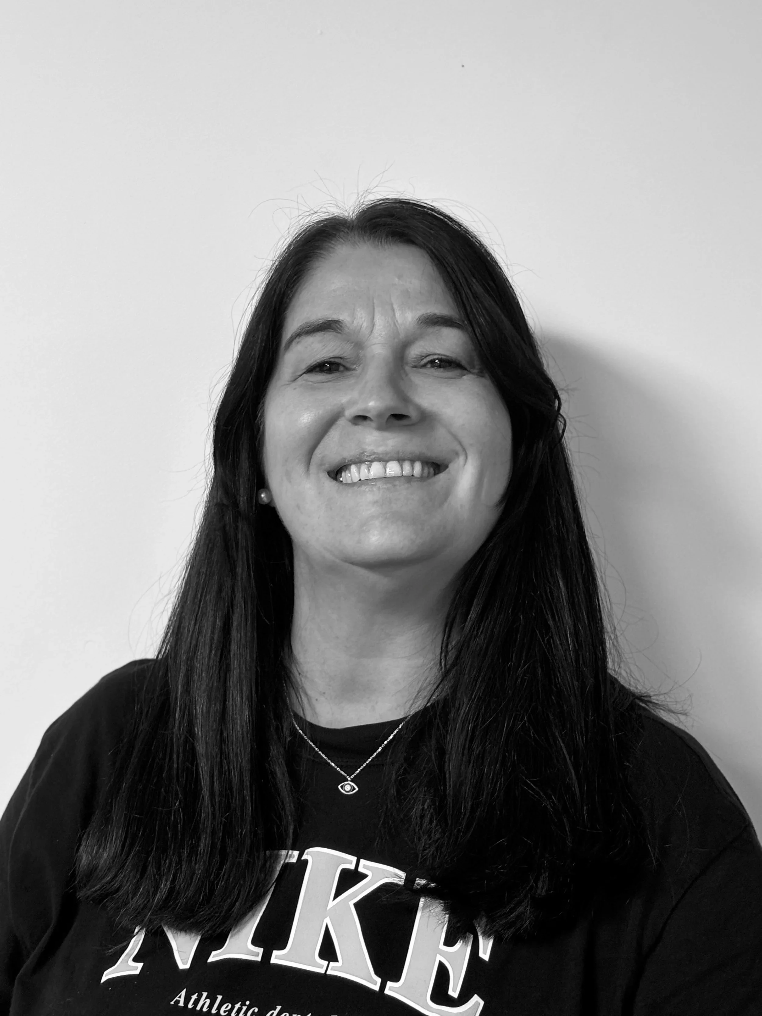 A smiling woman with long dark hair wearing a black Nike sweatshirt and a necklace with a circular pendant, standing against a plain white wall.