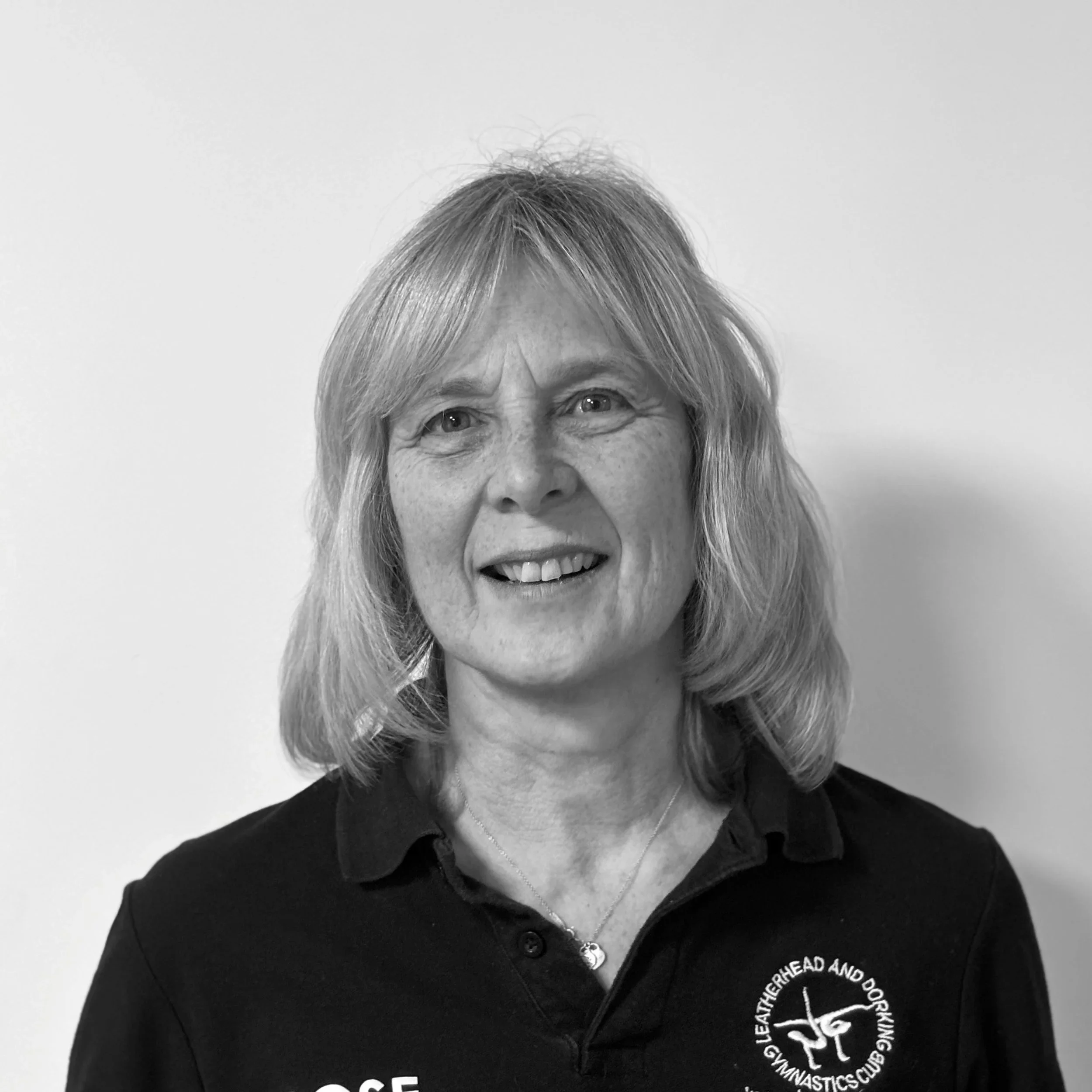 Close-up of a smiling woman with blonde hair wearing a black polo shirt with a logo on the left side that reads 'LEOTANG GYMNASTICS CLUB AND DAN' and a silver necklace, against a plain white background.