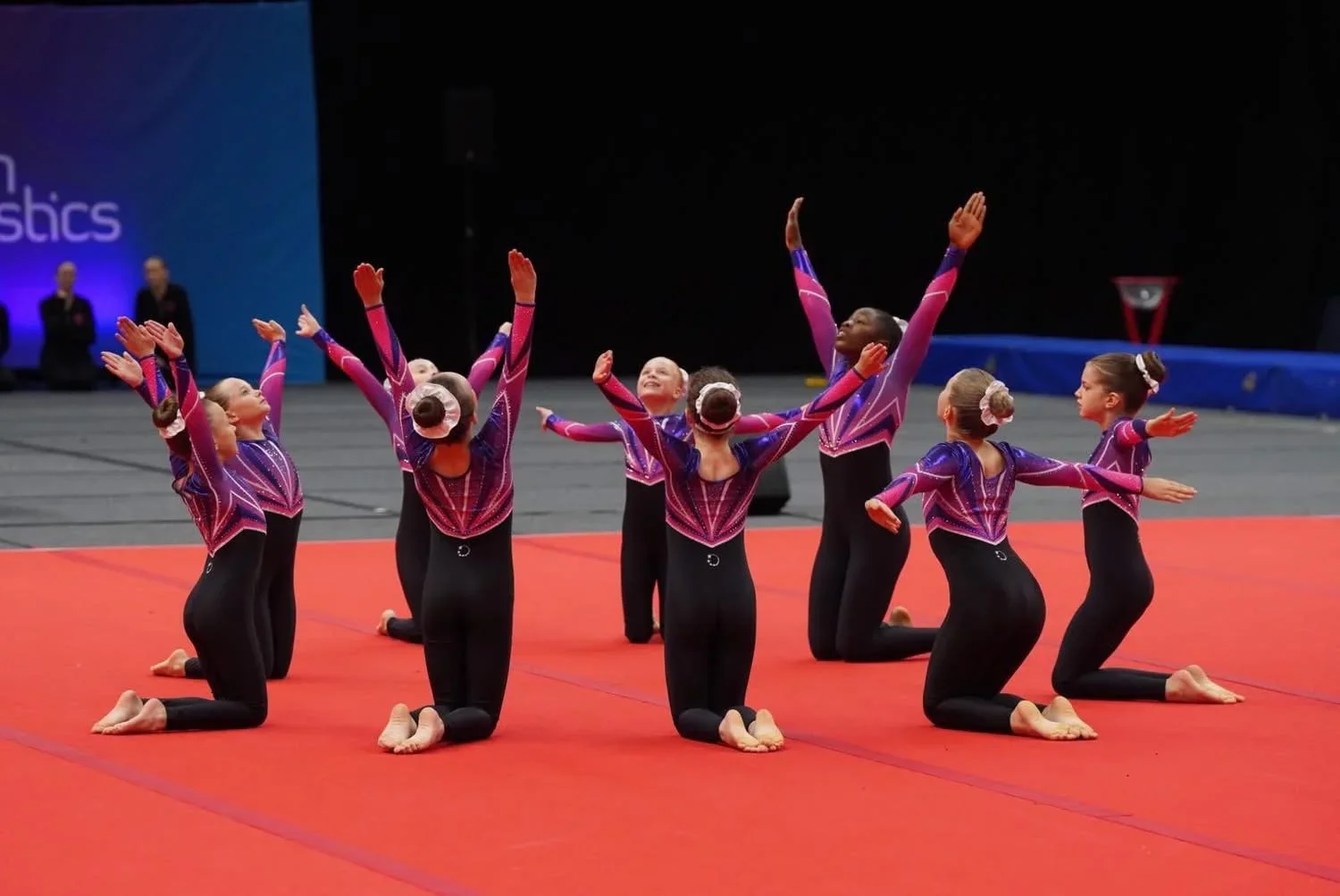 Young Team Gym gymnasts in purple leotards performing a synchronized routine on a red mat at a gymnastics competition.