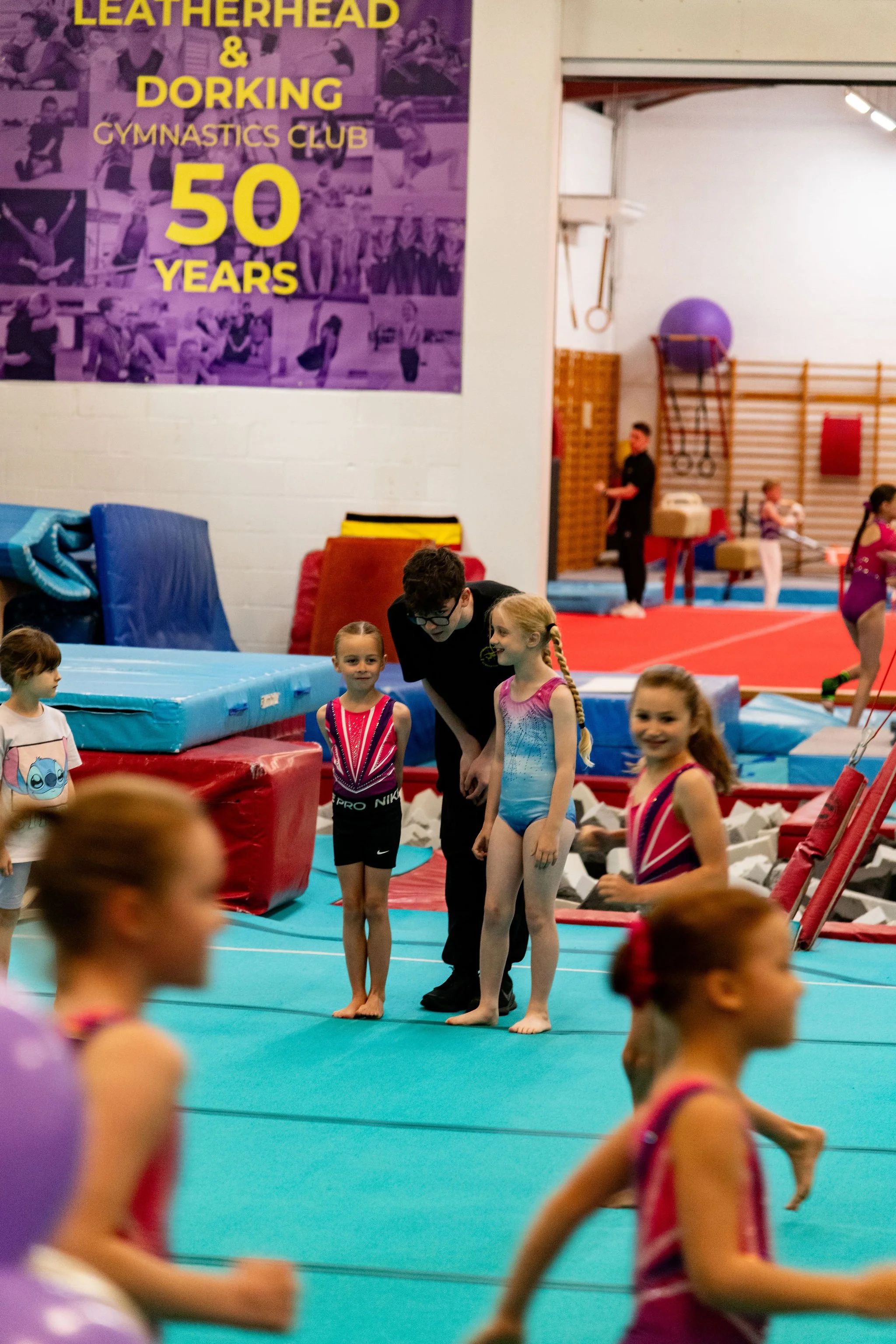 Children in a gymnastics gym, some standing on the mat, others running, with gym equipment and a purple exercise ball in the background, celebrating 50 years of a gymnastics club.