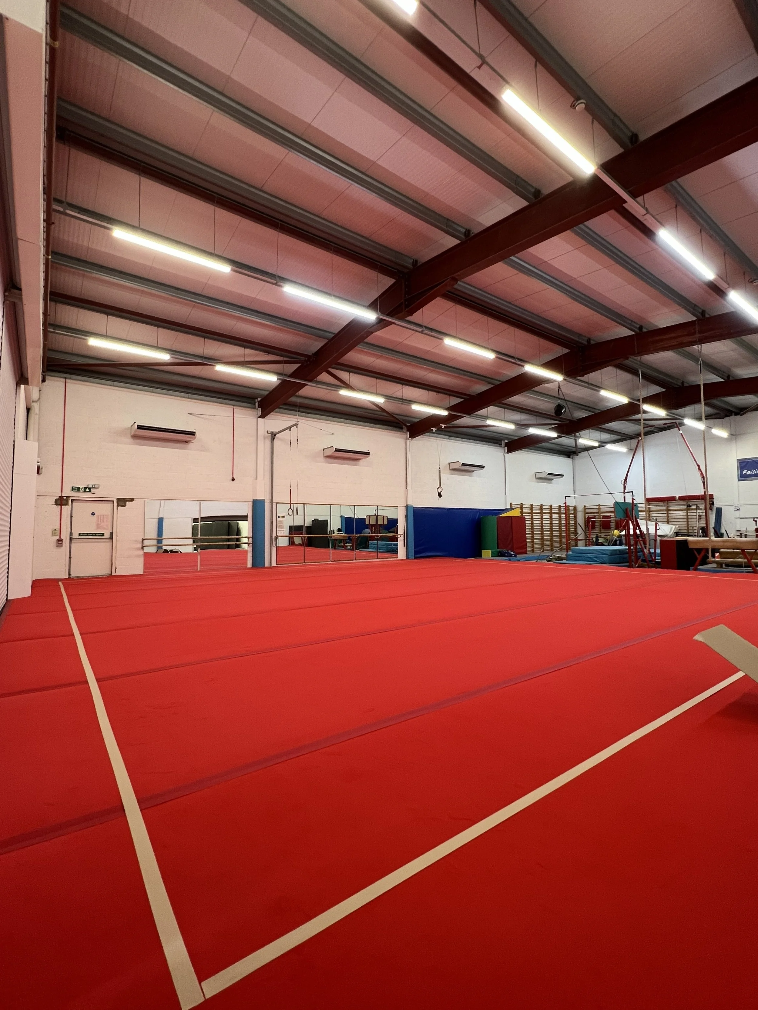Indoor sports facility with red gymnastics mats on the floor, mirrors on the wall, and various gym equipment in the background.