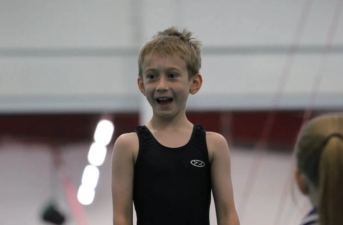 Young boy with blonde hair in a black athletic outfit smiling indoors.