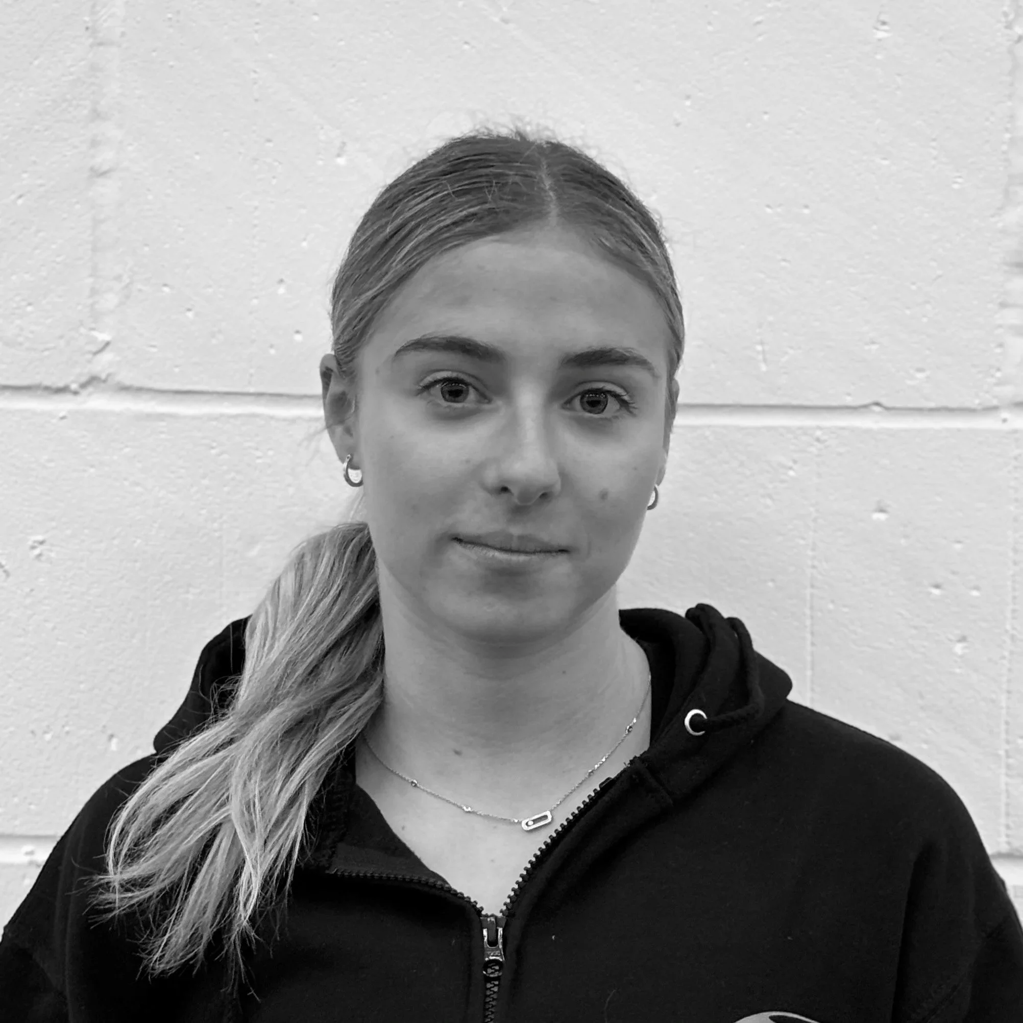 Close-up black and white photo of a young woman's face, highlighting her eyes, nose, and part of her forehead. She has light-colored hair pulled back and is wearing earrings and a necklace. She is dressed in a black zip-up hoodie.