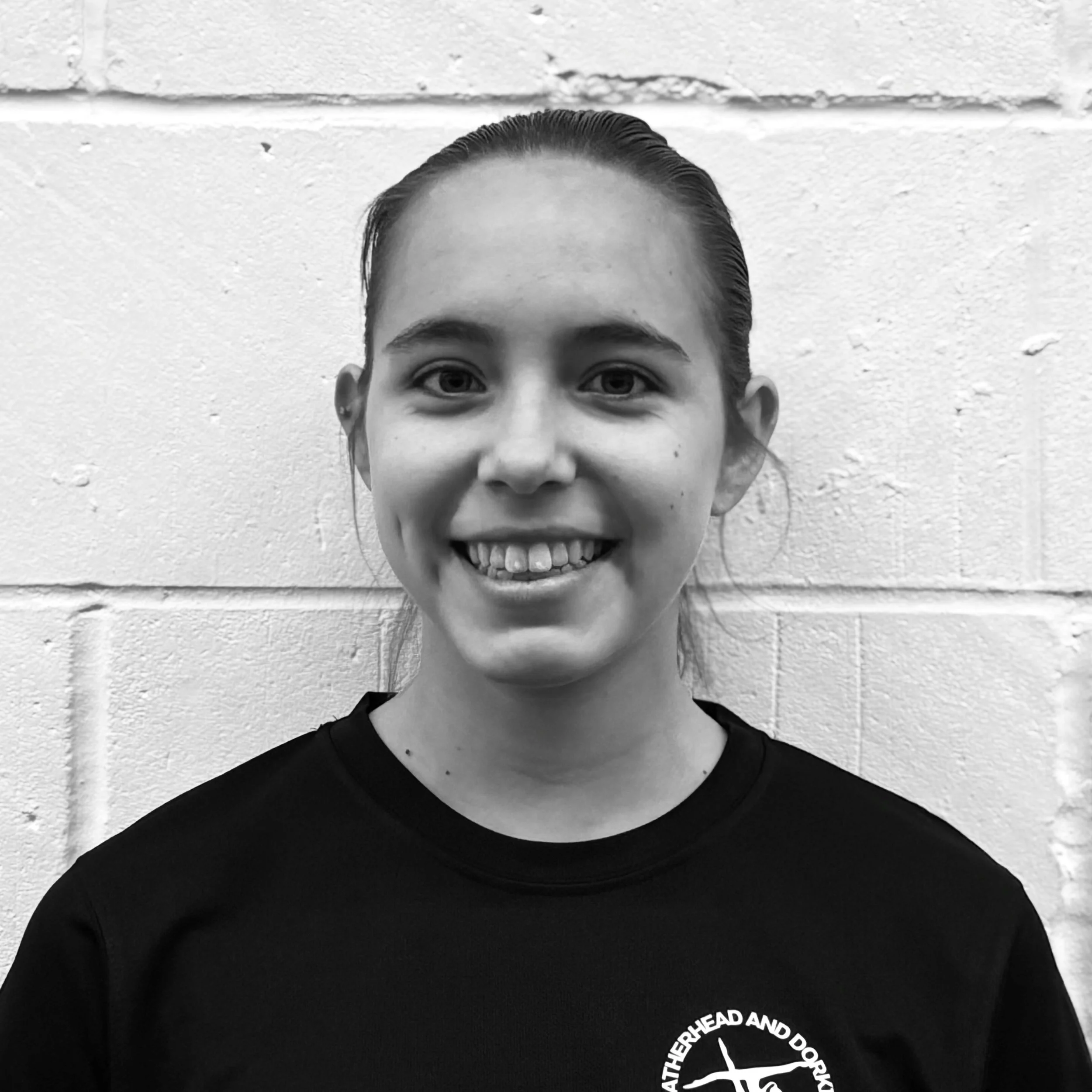 Close-up of a person with a partially visible face, smiling, against a brick wall, wearing a black T-shirt with a circular logo that reads 'ATHERHEAD AND DORSET'.