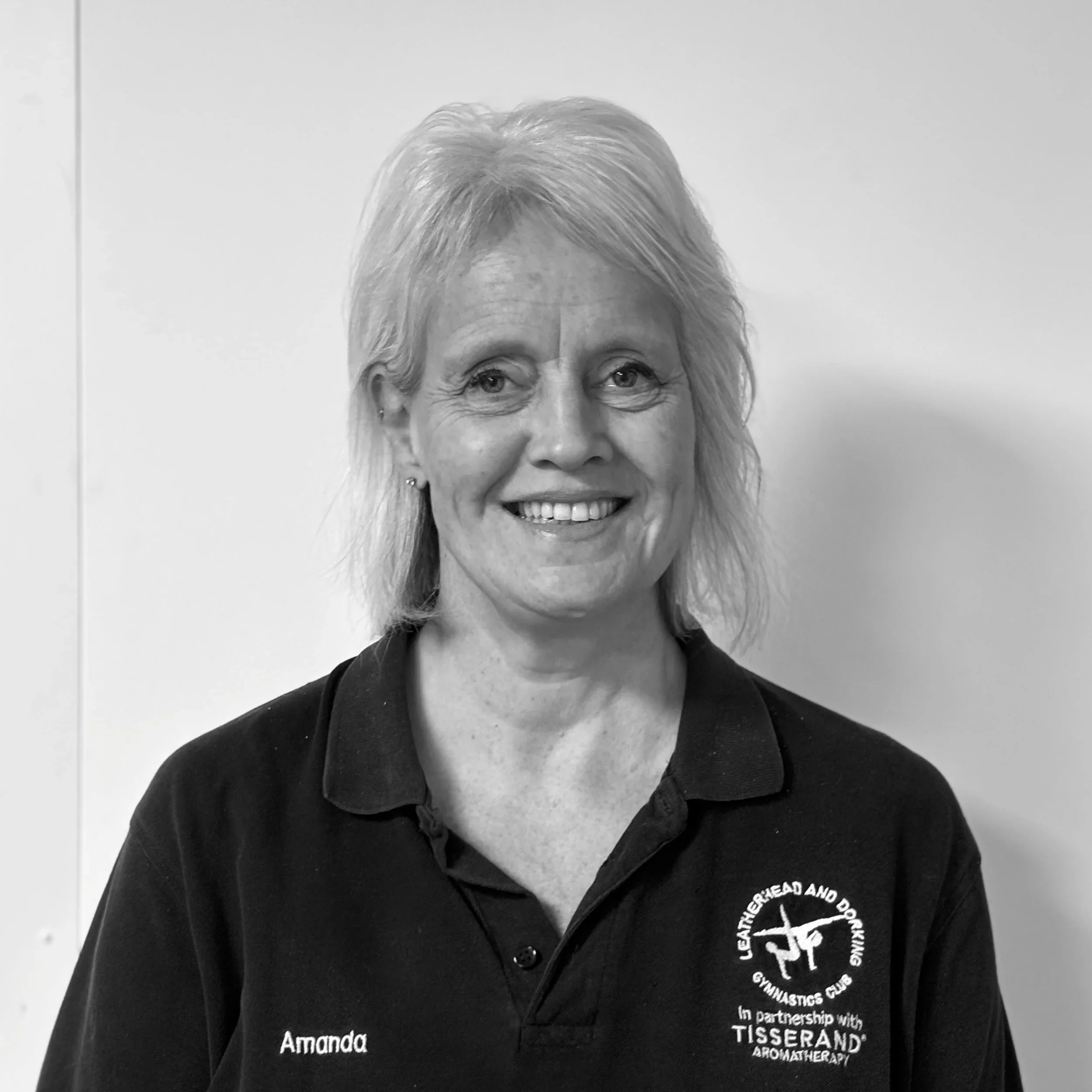 Close-up of a woman with short, light-colored hair, smiling, wearing a black shirt with embroidered text and logos, standing against a plain wall.