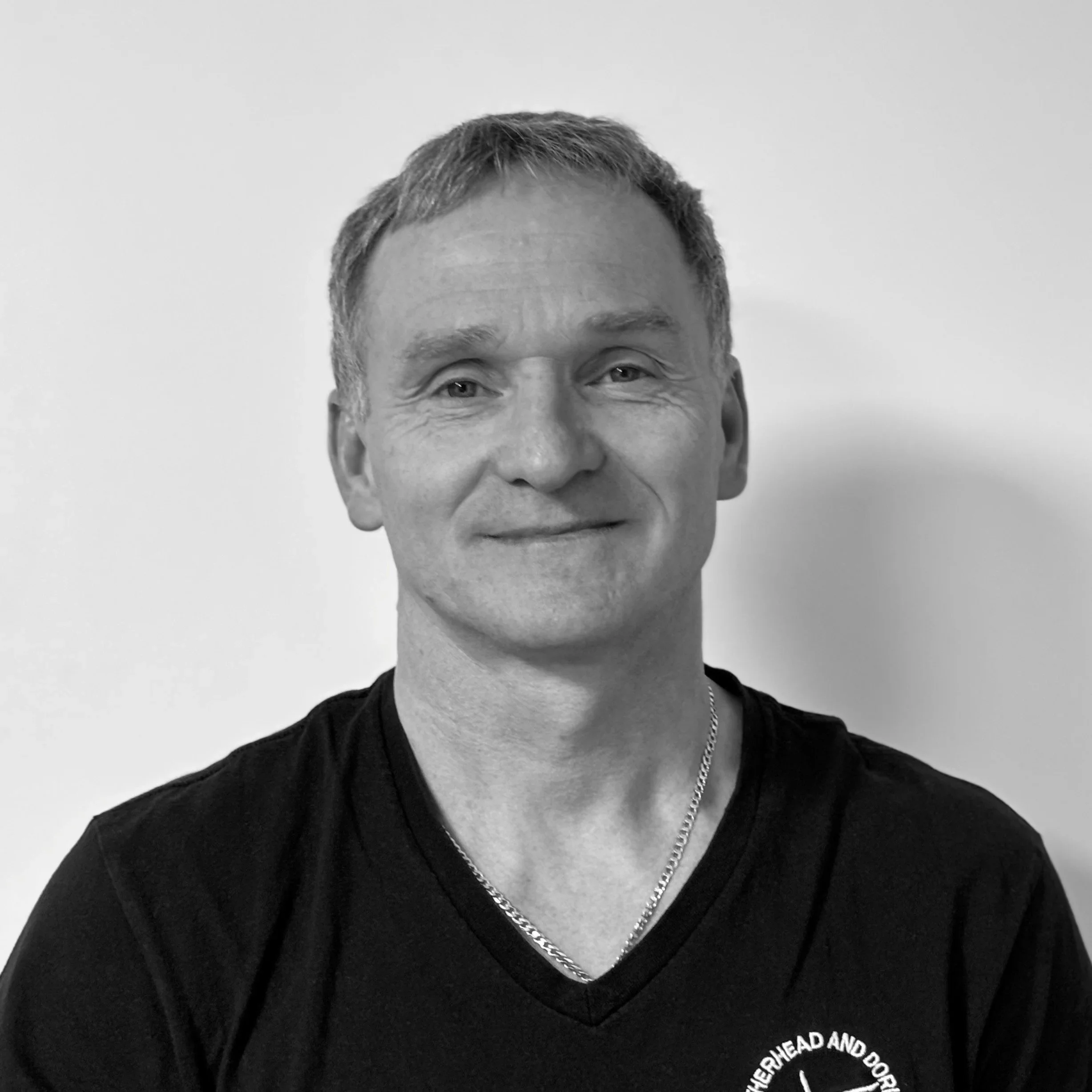 Close-up of a smiling man with short gray hair, wearing a black, V-neck shirt and a chain necklace, against a plain light background.