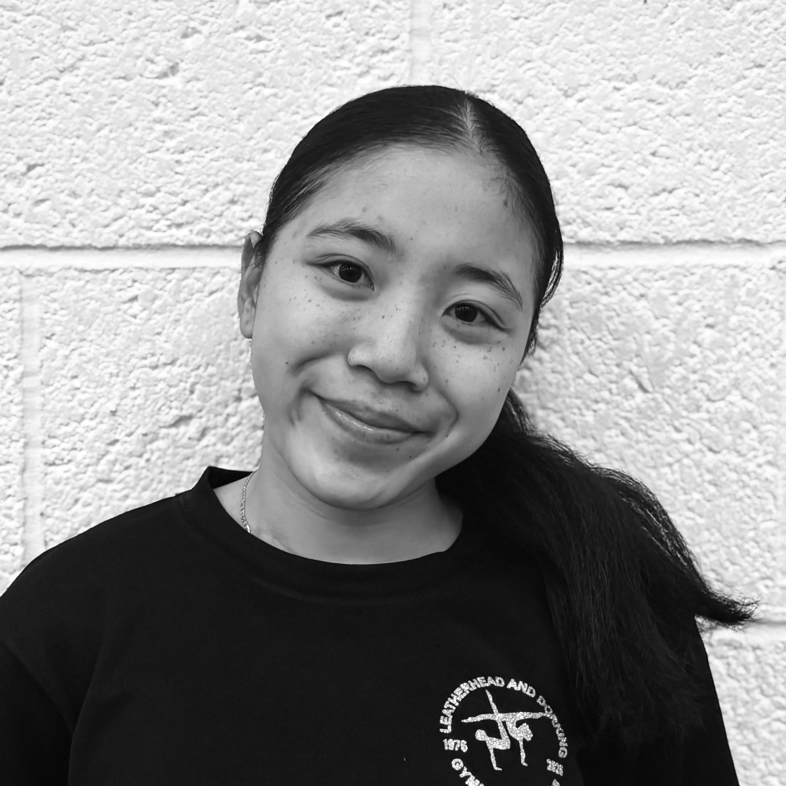 Close-up black-and-white photo of a young woman with dark hair, smiling, wearing a black t-shirt with a circular logo on the left side of her chest, standing in front of a textured wall.