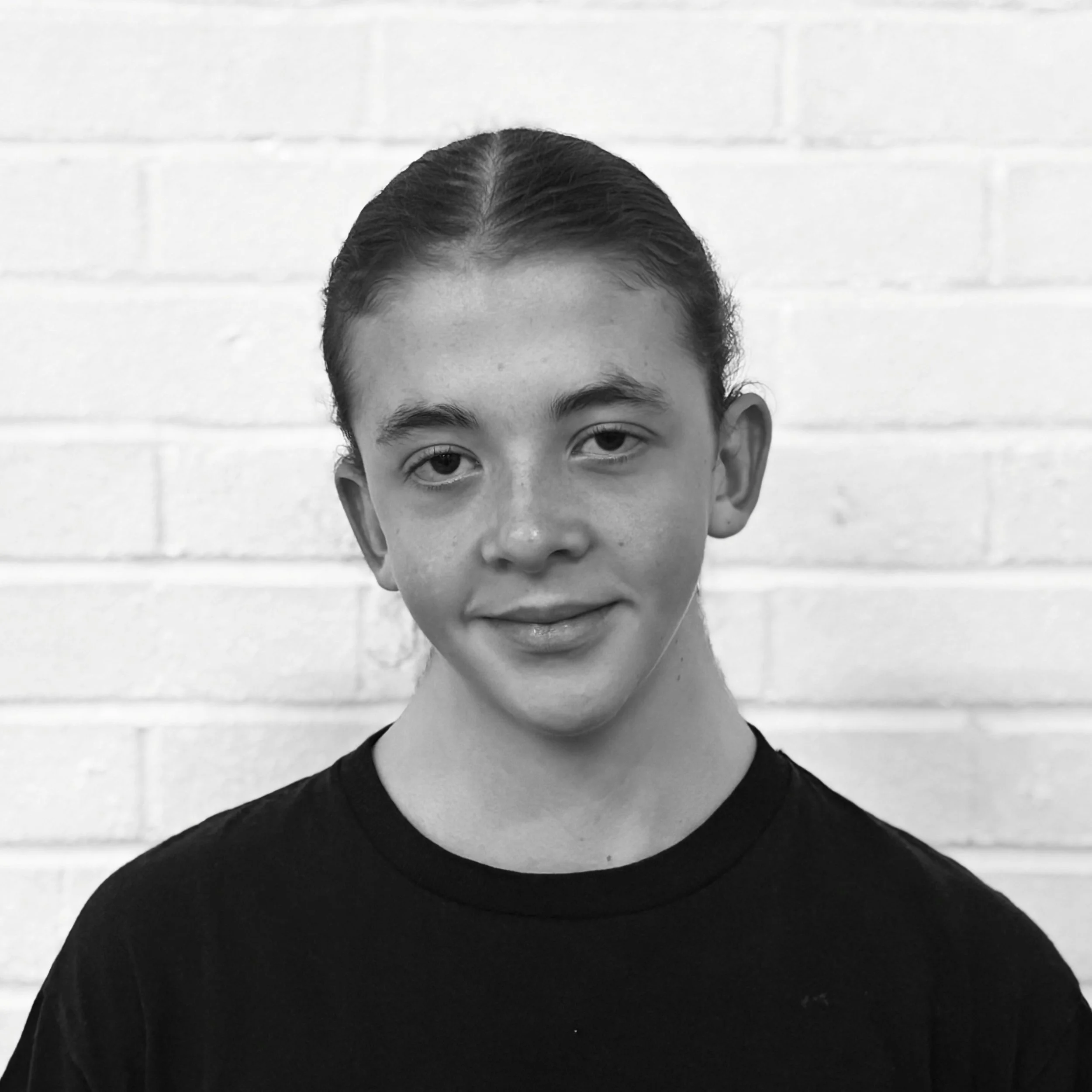 Black and white photo of a person with dark hair pulled back, wearing a black or dark-colored shirt, standing against a white brick wall.