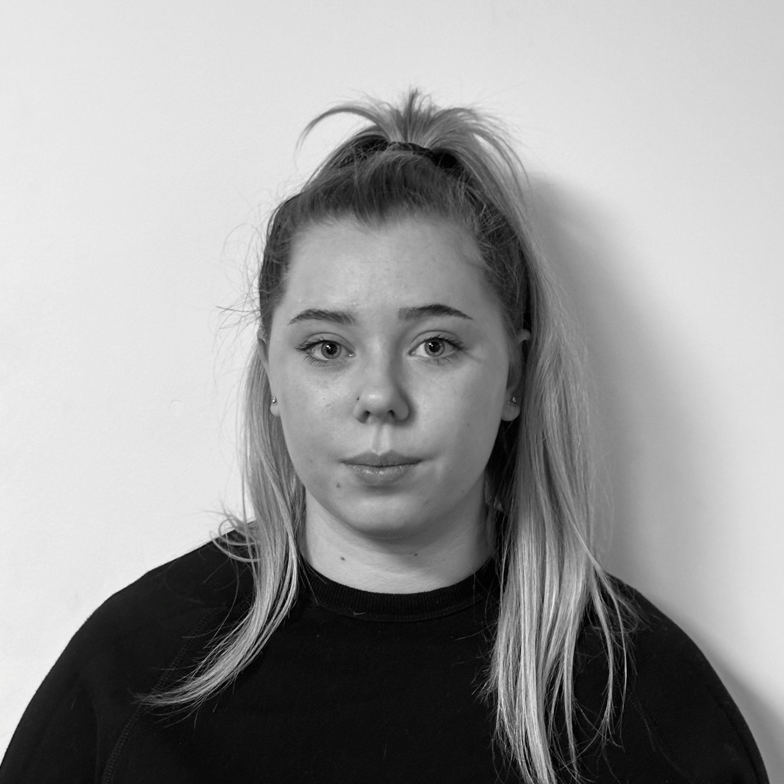 Black and white photograph of a woman with light-colored hair styled in a high ponytail, wearing a black top, standing against a plain, light-colored wall. Only the upper part of her face including eyes, eyebrows, nose, and mouth is visible.