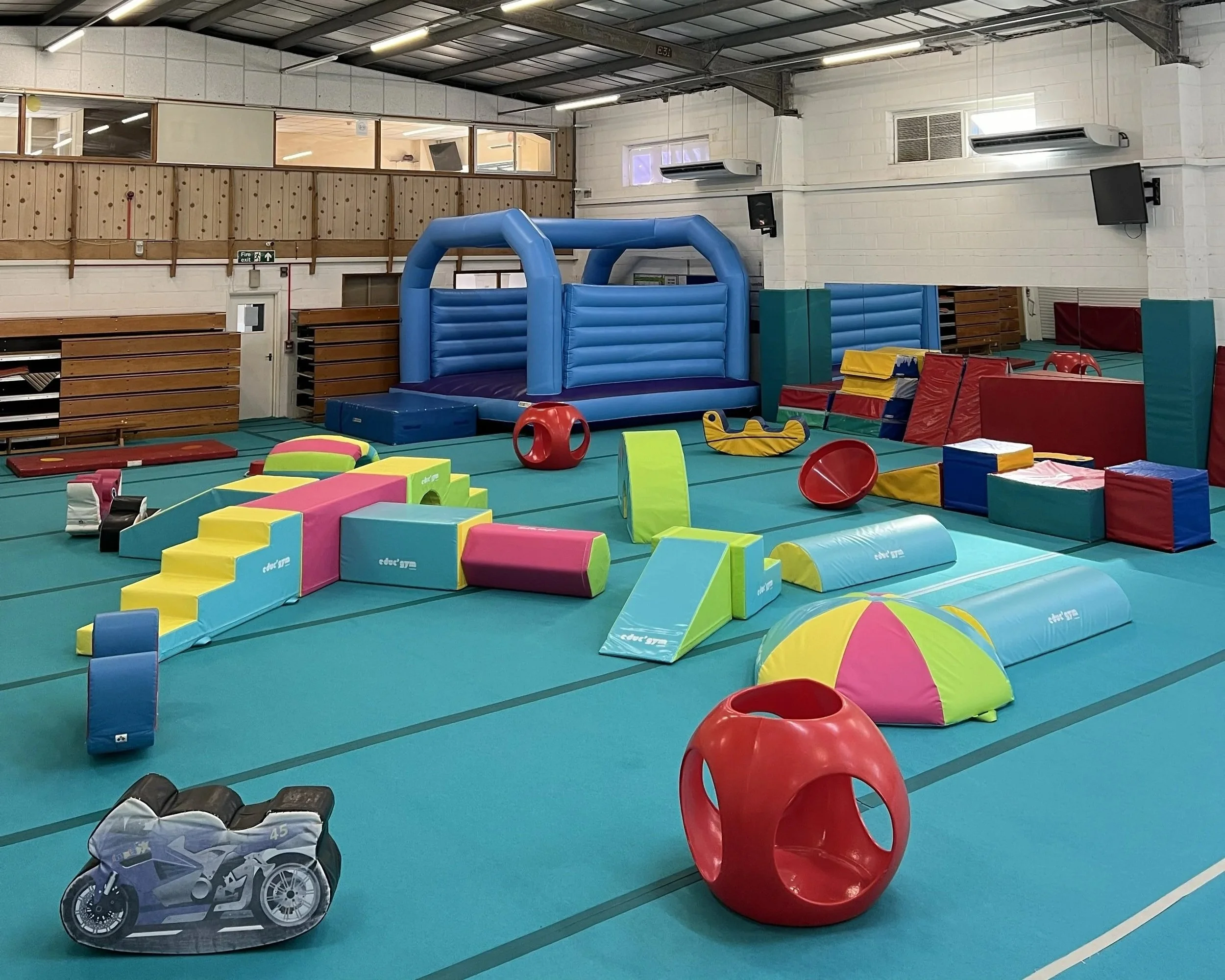 Indoor gymnasium with colorful soft play equipment, including a large blue bounce house, foam blocks, a red plastic chair, and a toy car. The background shows a wooden balcony, air conditioning units, and a TV mounted on the white brick wall.
