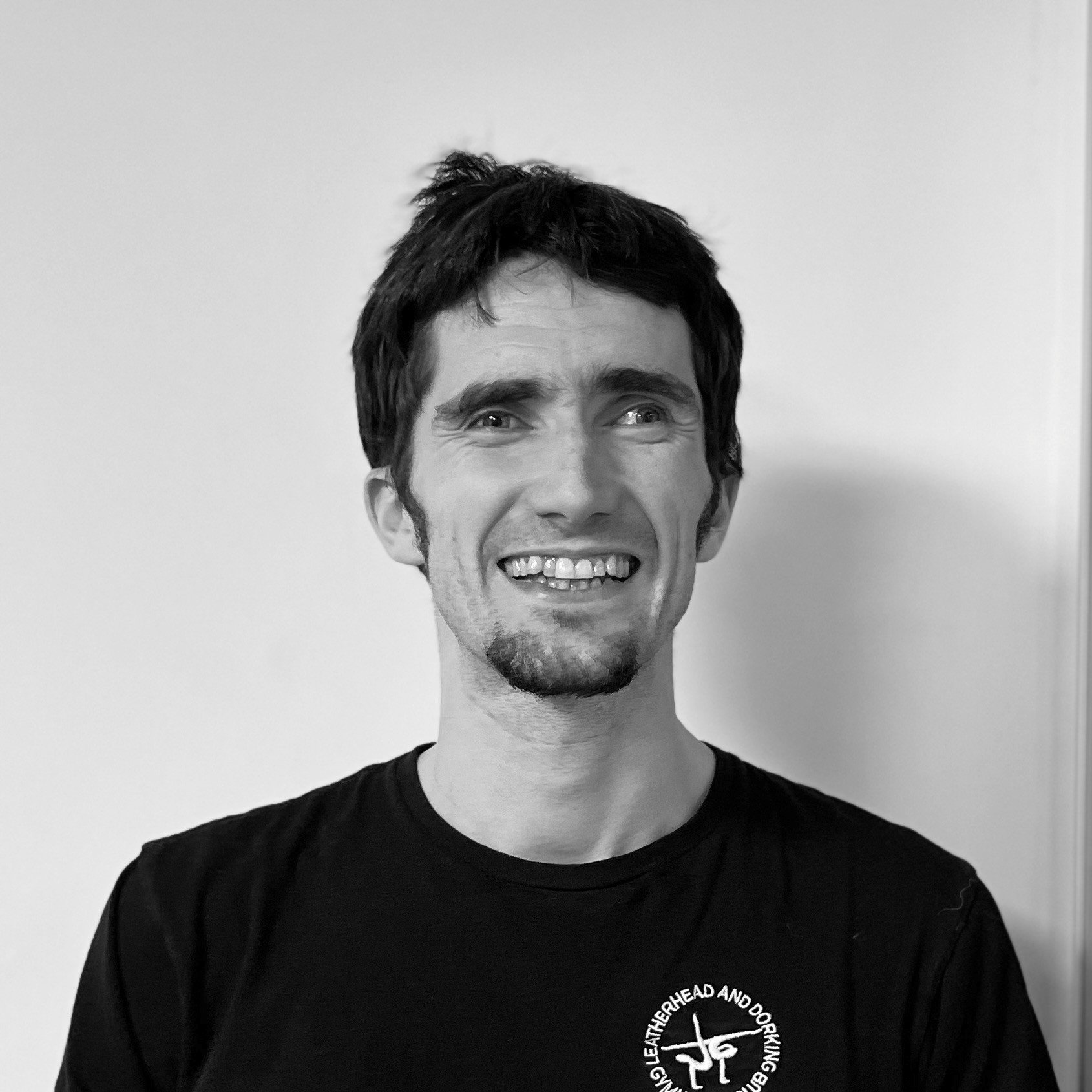 Black and white photo of a young man with short dark hair, smiling with teeth showing, wearing a black T-shirt with a circular logo on the left side.
