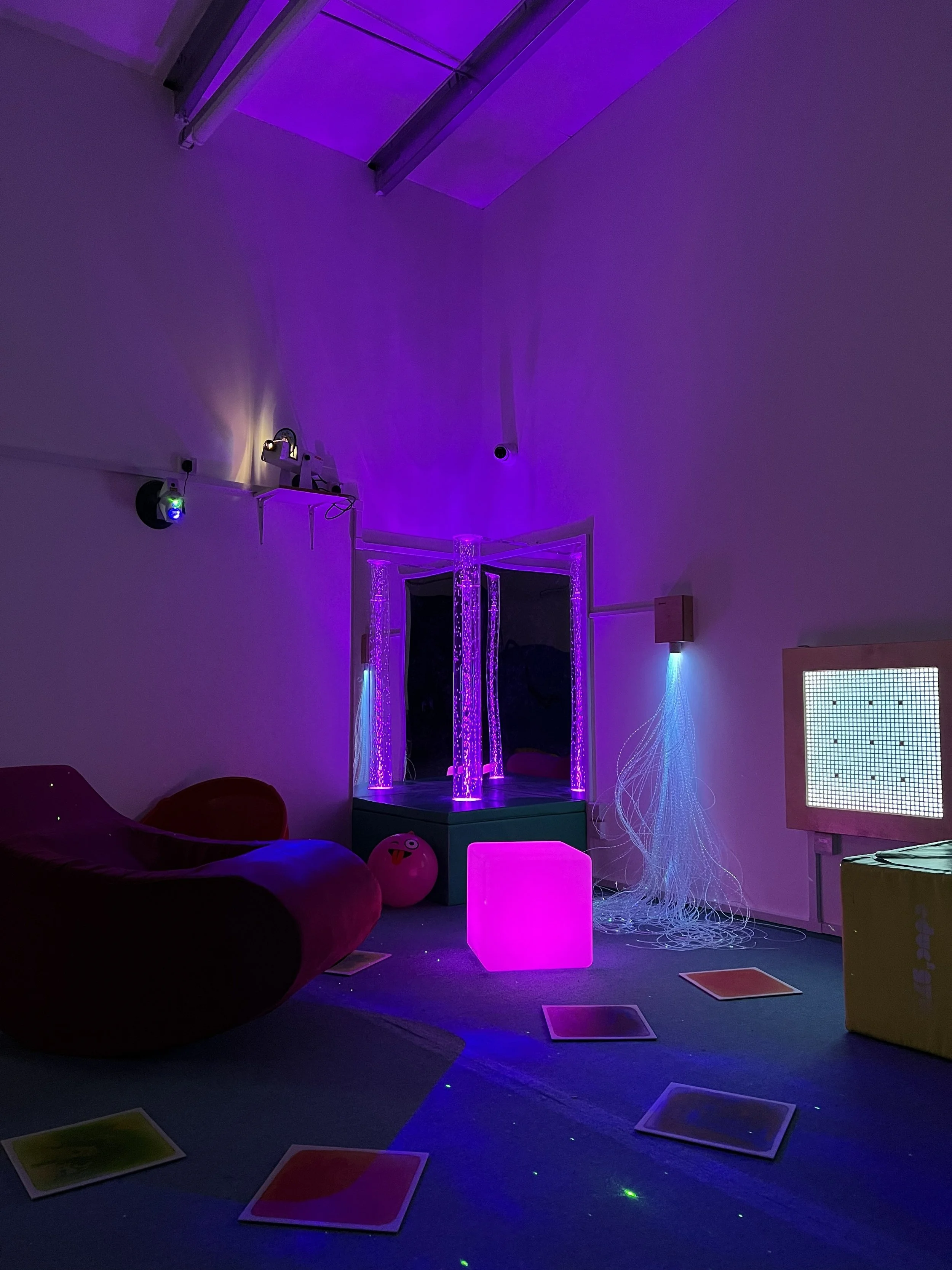 Room with purple and blue LED lighting, a hanging mirror, a pile of plush toys, a cube-shaped light, scattered colorful cards on the floor, and various decorations.