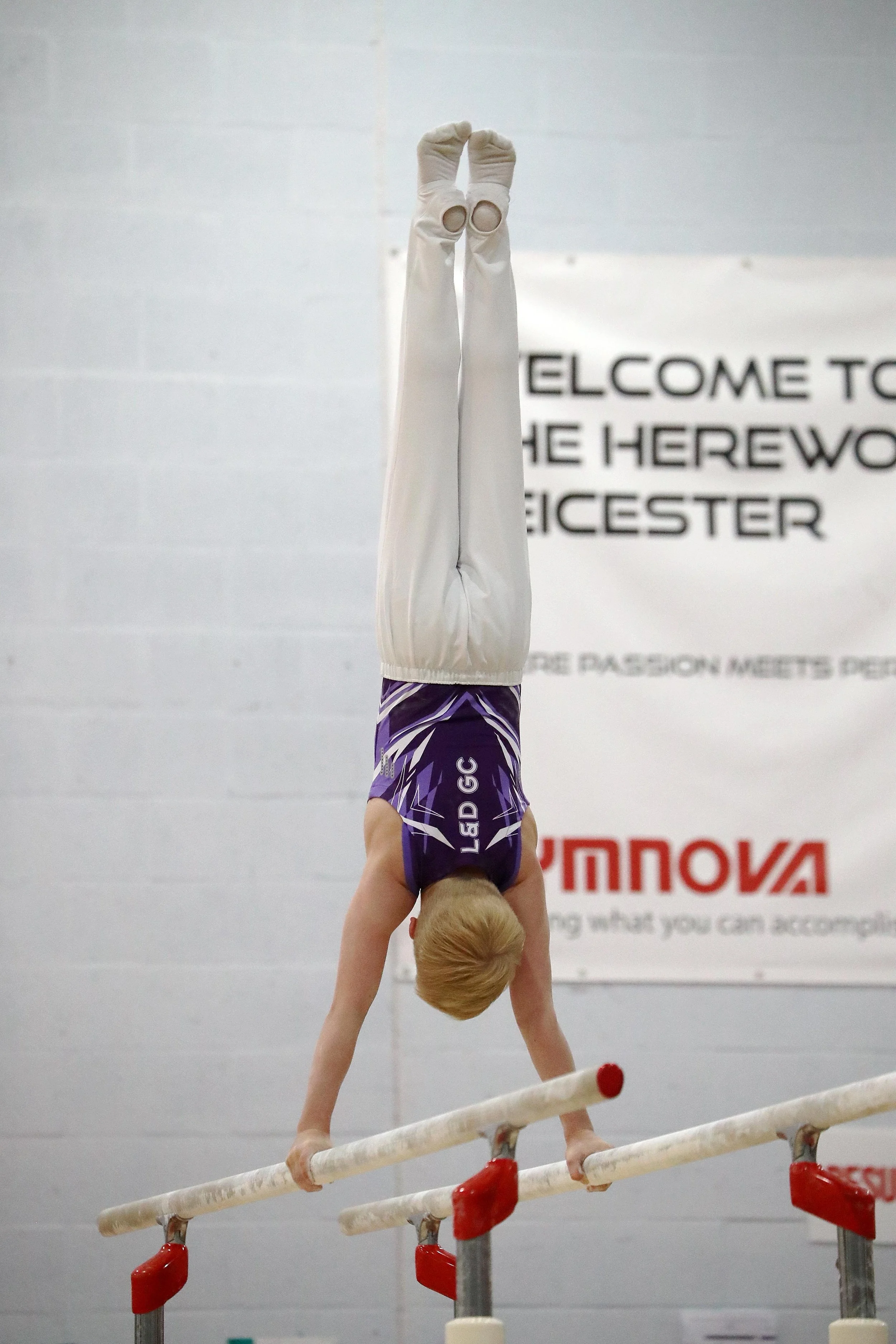 A gymnast in a purple and white leotard is performing a handstand on a balance beam in an indoor gymnastics facility with a white wall and black and white signage in the background.